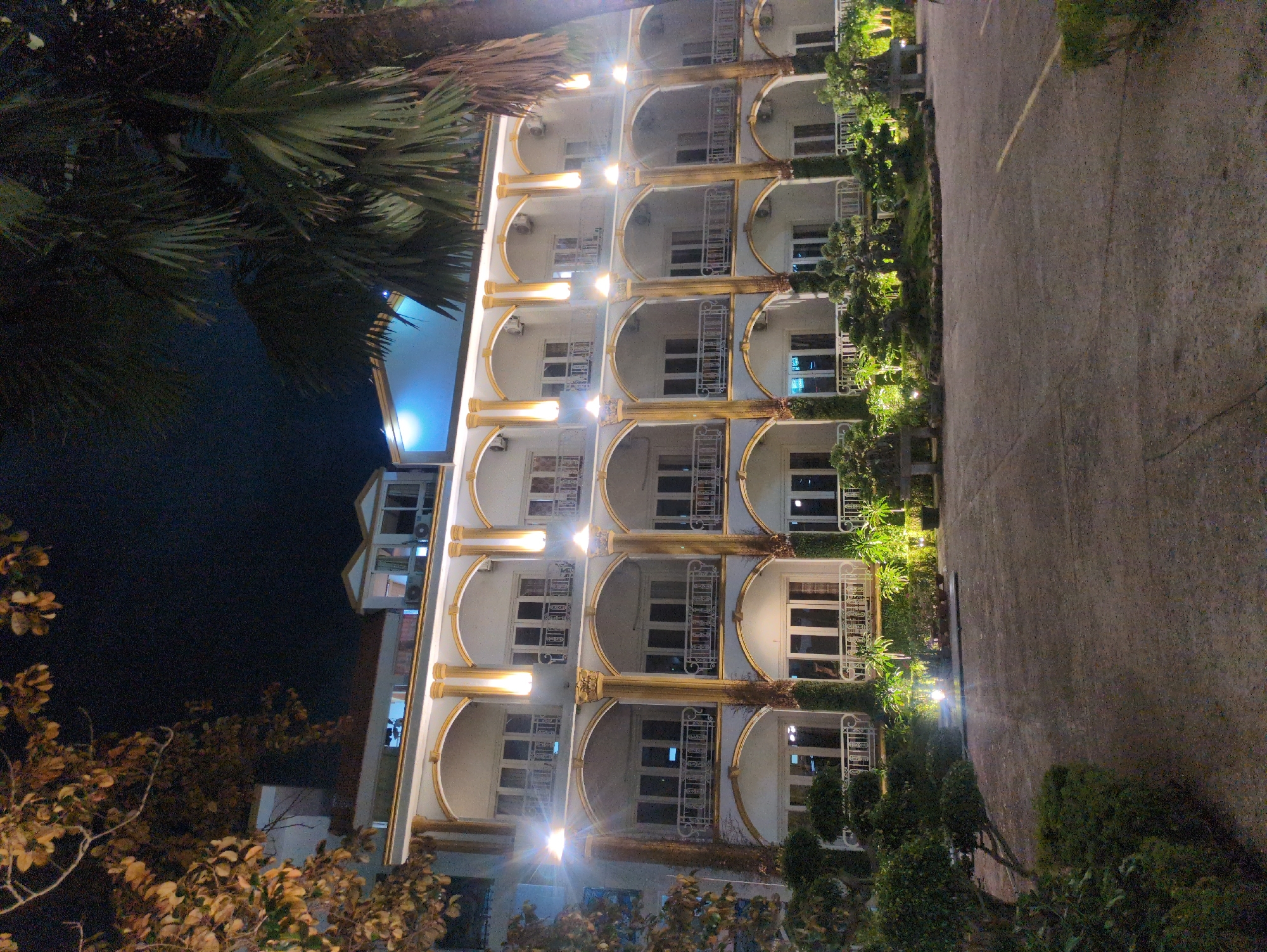 The image shows a multi-story hotel with balconies in Phú Quốc, Vietnam, at night. The building has a white facade with gold accents and arched balconies. Each balcony has a white railing. Some balconies have curtains drawn. There are lights positioned on the building facade that illuminate the balconies and the walkway. The surrounding environment includes palm trees, shrubs, and trees, and an adjacent paved area. There are no visible people in the scene.