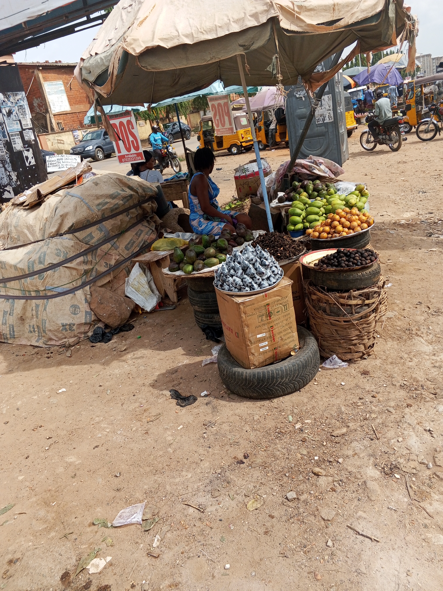 The image depicts a street market stall in Abuja, Nigeria. A woman sits under a large, worn umbrella, likely selling various fruits and nuts. The stall is set up on a dirt patch, and merchandise is displayed on tables and elevated platforms made of stacked tires, a cardboard box, and a woven basket. The items on display include mangoes, avocados, nuts (some wrapped in small foil packages), and other produce. Behind the stall, there are bags of materials stacked. There is activity in the background, with motorcycles and "keke napep" auto-rickshaws moving along the road. There is a POS sign, and other signs visible on the buildings. Debris is on the ground surrounding the stall.