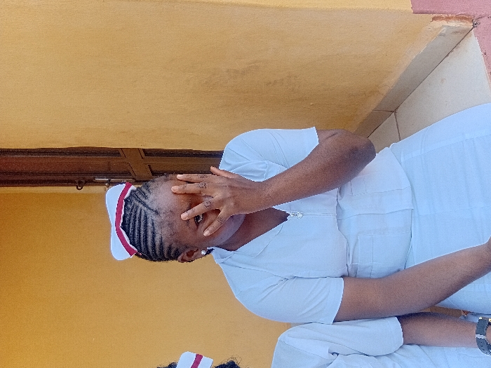 An adult female of African descent is depicted in Ejule, Nigeria. She is wearing a white short-sleeved uniform with a V-neck and a metallic zipper pull, and a matching white skirt. A white cap with a red band is worn on her head, over dark cornrowed hair. Her right hand covers the lower half of her face, partially obscuring her nose and mouth, with her left eye looking directly at the camera. A small earring is visible in her left ear. To her immediate left, the partial figure of another individual, also in a white uniform, is visible, including a left wrist adorned with a dark-strapped watch. The background consists of a plain, warm yellow-orange wall. A dark brown wooden door frame is partially visible vertically on the right side of the frame. The subjects appear to be seated, with a tiled floor visible in the lower foreground, featuring light-colored tiles transitioning to a reddish-brown base.