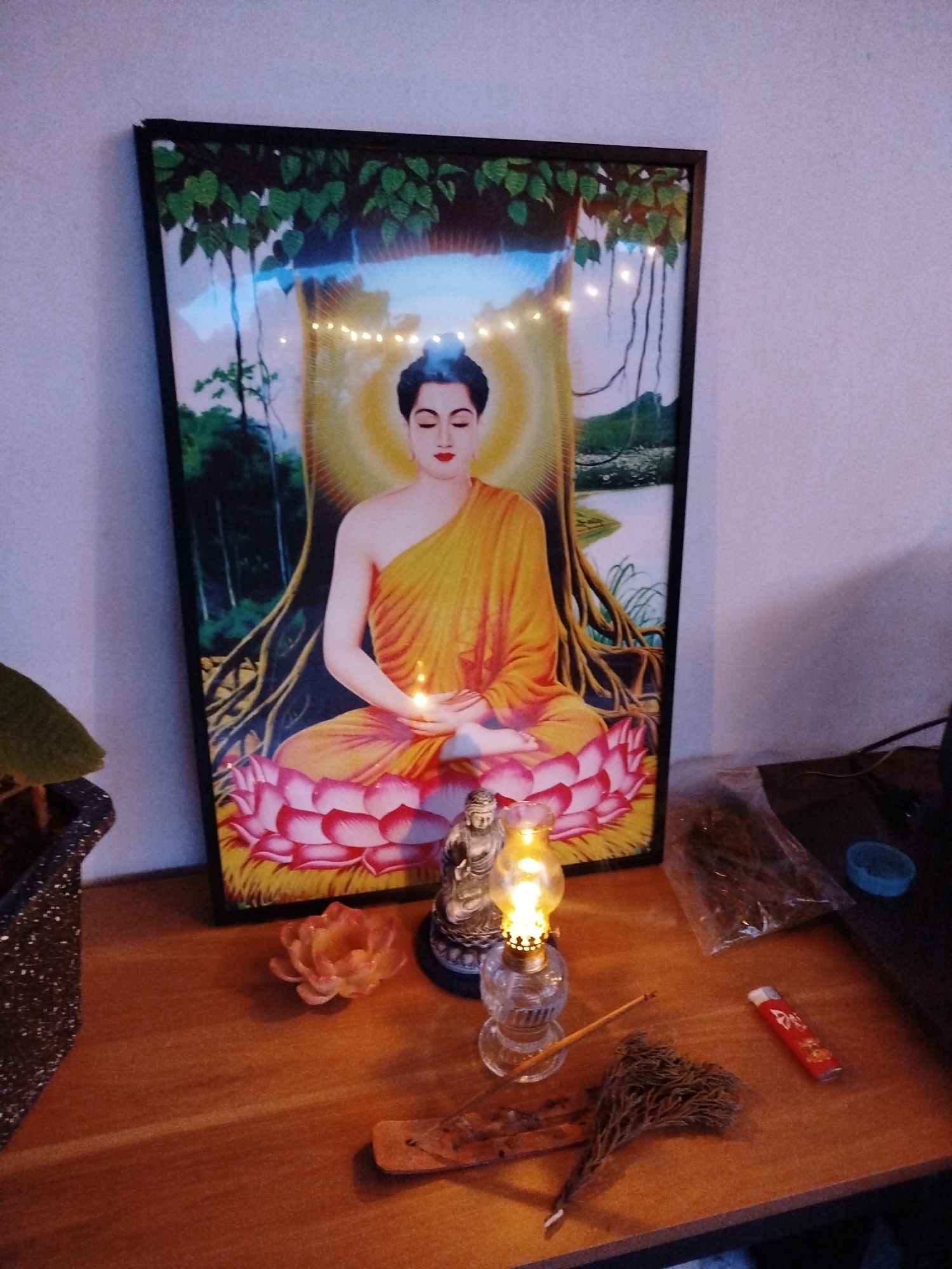 The image depicts a devotional altar set up in what appears to be a home environment in Lạc Dương, Vietnam. The central subject is a framed picture of the Buddha meditating under a Bodhi tree, with a glowing aura and a lotus flower base. String lights are draped over the top of the frame, adding a decorative touch.
In front of the picture, a small Buddha statue sits beside a miniature oil lamp, its flame casting a warm glow. A single unlit incense stick rests on a wooden stand, with a bundle of dried herbs or incense material nearby. A small, decorative lotus flower sits to the left of the incense. To the right, a red lighter and a clear plastic bag filled with dried plant material are visible. A small blue cap or container is also present. A plant in a textured pot is partially visible on the far left.
The lighting suggests it might be evening or a dimly lit indoor space, creating a serene and contemplative atmosphere. The overall scene evokes a sense of spirituality and personal devotion. No visible text is present in the image.
