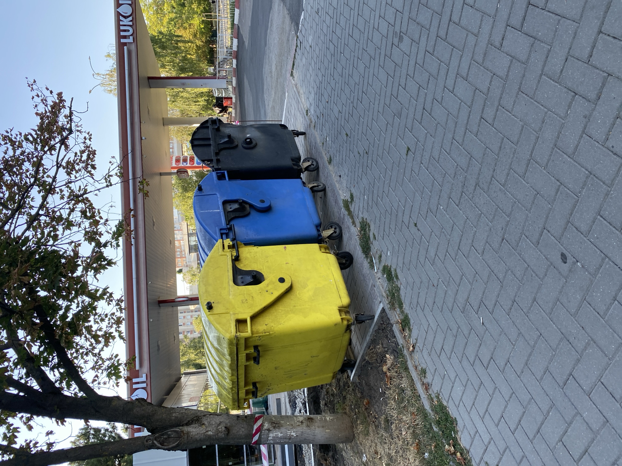 The image depicts a scene at a Lukoil gas station in Chișinău, Moldova. In the foreground, three large, wheeled trash bins are lined up on a paved sidewalk. The leftmost bin is bright yellow, followed by a blue bin, and then a black bin. A tree with sparse leaves stands to the left of the bins, with a striped pole next to its base. Behind the bins, the gas station canopy is visible, with "LUKOIL" signage prominently displayed. Gas pumps are partially visible under the canopy, along with a price display. The background shows buildings and trees, indicating an urban setting. The sky is clear and blue. The overall impression is a typical street scene with commercial and waste management infrastructure.