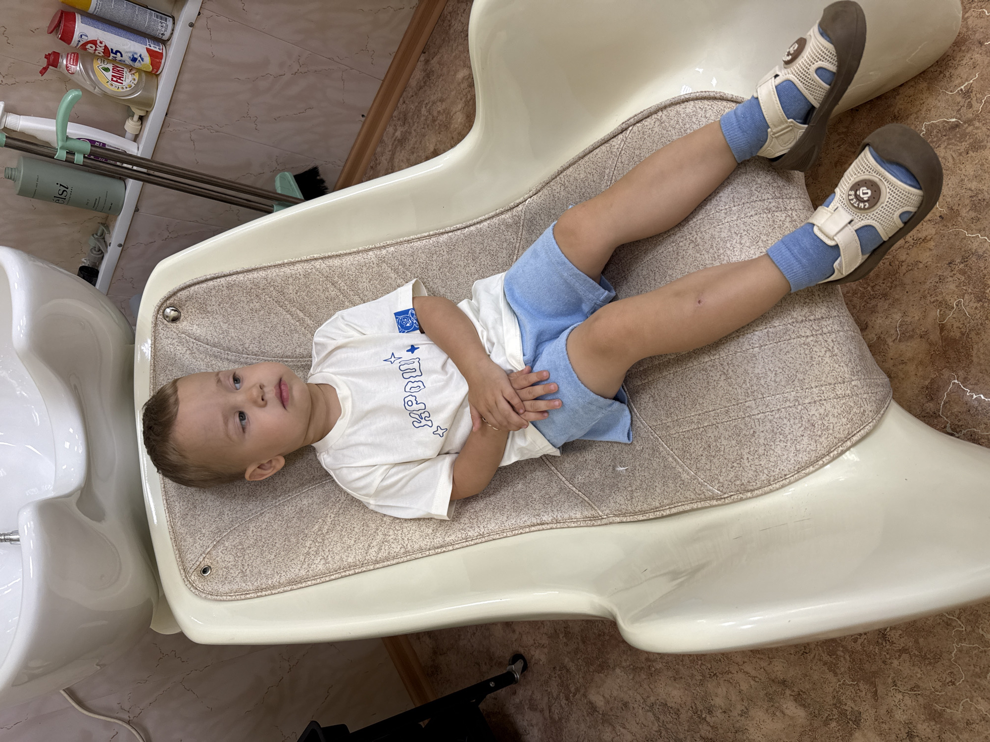 A young boy with light brown hair and fair skin is sitting in a cream-colored shampoo chair in what appears to be a hair salon in Rusanovo, Russia. He is wearing a white t-shirt with blue text and graphics, and light blue shorts. He has on blue socks and light brown and cream-colored sandals. His hands are clasped in his lap. To the left of the chair, a portion of a black rolling cart is visible. Behind the boy, on a shelf mounted on a light pink tiled wall, are several bottles of hair products, a broom, and other cleaning supplies. A white sink is positioned above the shampoo chair. The floor is tiled with a brown and beige pattern.