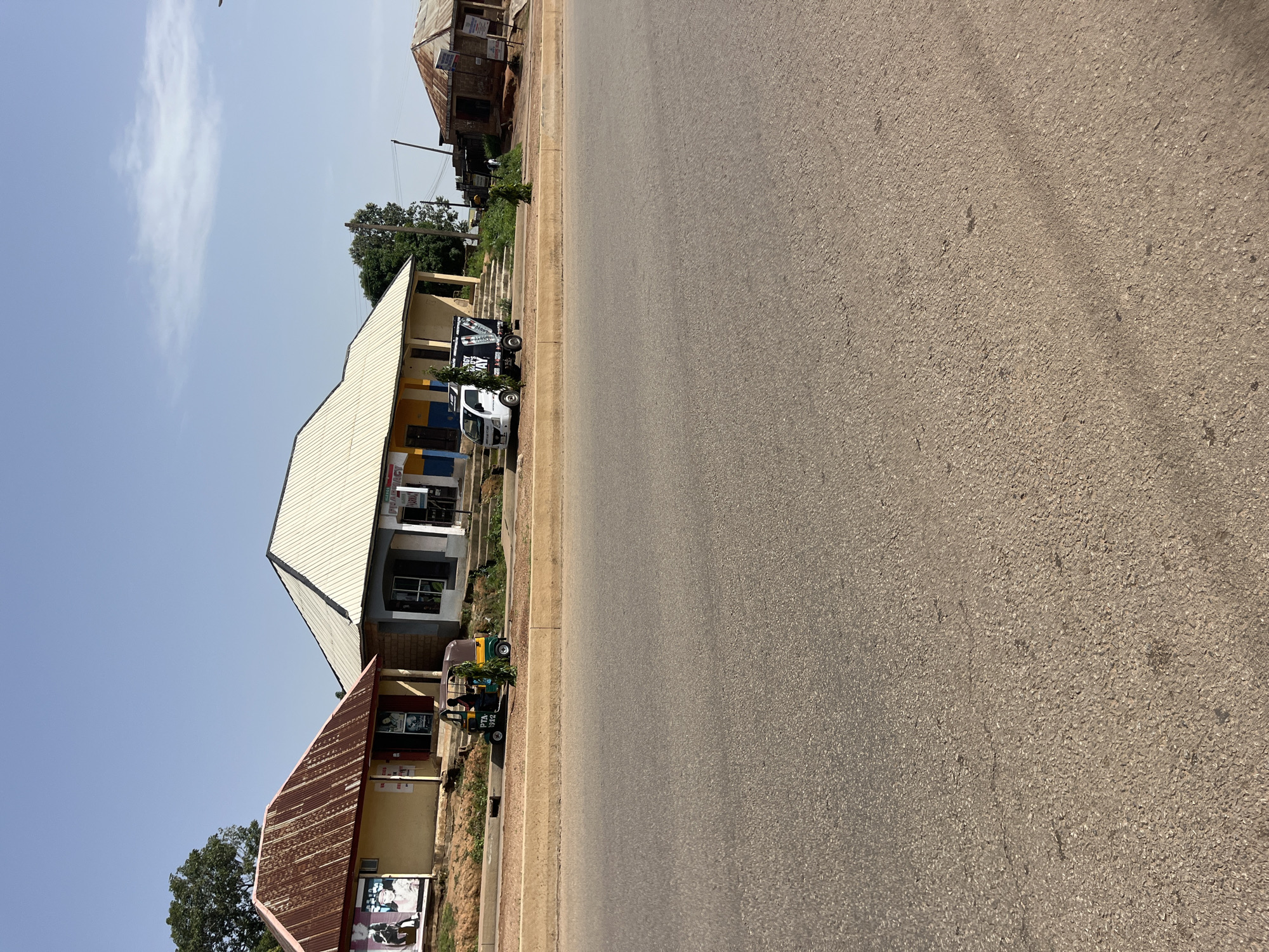 The image captures a street scene in Bukuru, Nigeria, on a clear day with a bright sky. In the mid-ground, a row of buildings lines the side of a paved road. The first building on the left has a corrugated metal roof and features several posters on its facade, one depicting two individuals. Adjacent to it is a larger building with a lighter, possibly aluminum, sloped roof, displaying signs for a pharmacy. A white truck with a dark-colored advertisement on its side is parked in front of this building. To the right of this structure, another small, weathered building with a corrugated roof is visible.
In the foreground, the road surface is asphalt, appearing worn in places. A beige concrete barrier separates the road from the area where the buildings and vehicles are situated. Parked along this barrier is a three-wheeled motorized vehicle, commonly known as a "Keke Napep" or "Marwa," with a driver seated inside and a passenger or cargo area behind. A small, green leafy plant is positioned next to this vehicle. The environment suggests a commercial or mixed-use area within the city.