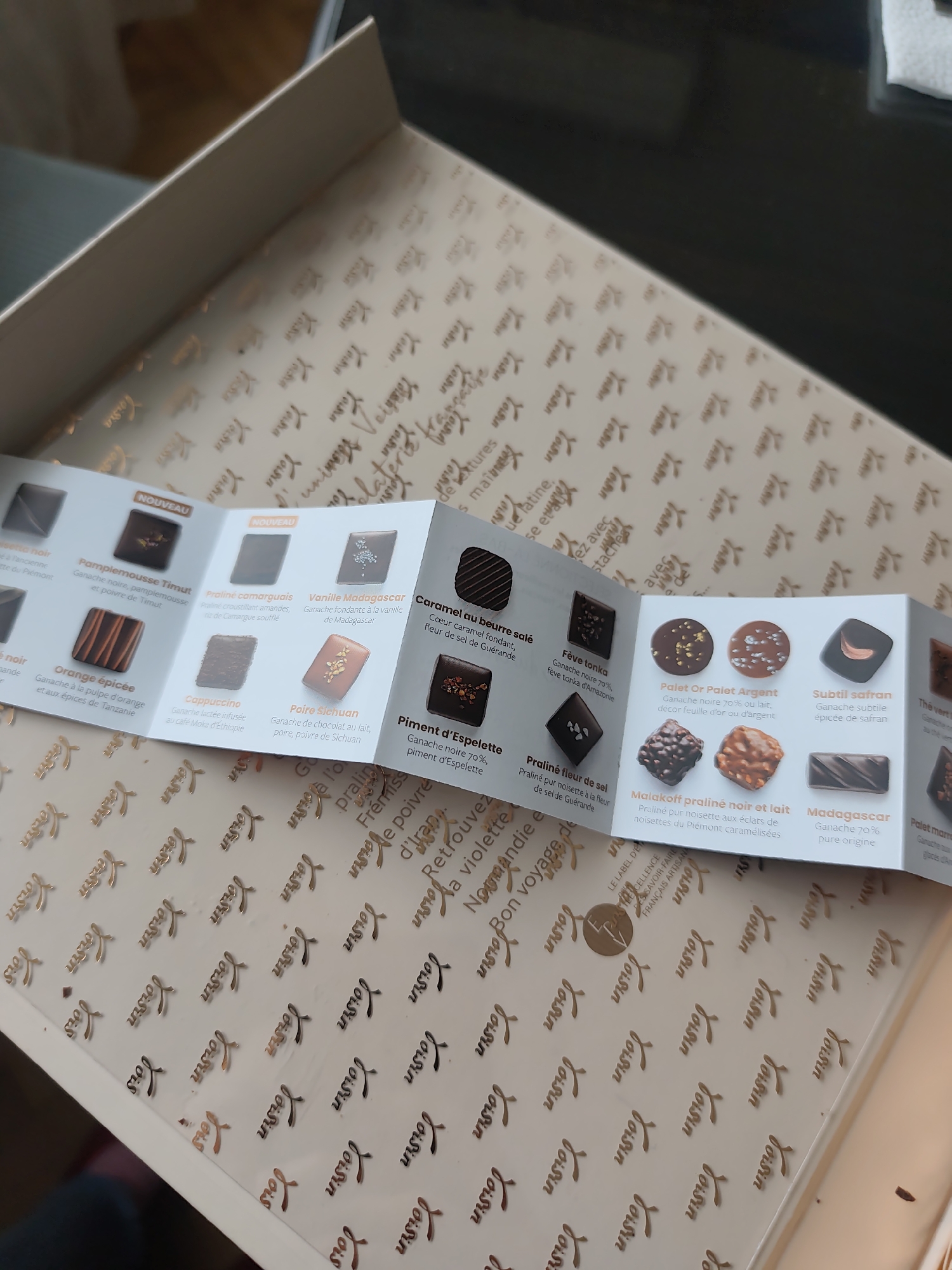 The image shows an open box of "Voisin" chocolates, with a folded information sheet displaying pictures and descriptions of the different chocolate varieties. The sheet includes details such as flavor profiles, ingredients, and origin of ingredients, such as the "Pamplemousse Timut" with ganache noire and Timut pepper, or the "Caramel au beurre salé" featuring salted butter caramel and fleur de sel from Guérande. The box itself is patterned with the "Voisin" logo in a repetitive design. The overall setting appears to be indoors.