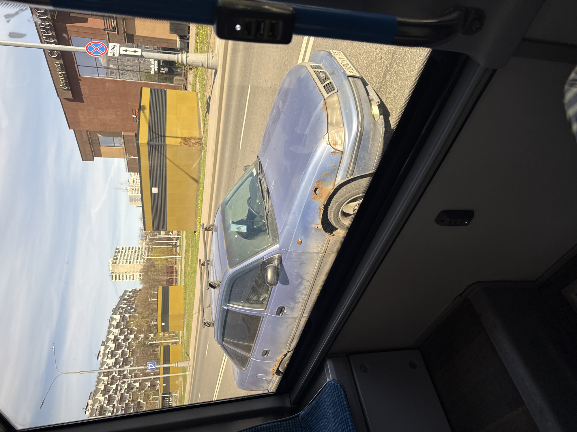The image captures a street scene in Moscow, Russia, viewed from inside a moving vehicle, likely public transport, during the daytime with clear, sunny weather. The primary subject is a heavily rusted blue car, possibly a Daewoo Nexia, parked alongside the road. The car exhibits significant rust on its front fenders, wheel arches, and lower body panels. It is equipped with roof racks. Its Russian license plate, visible in one crop, reads "P380VY 197", with "197" confirming the Moscow region.
To the left of the car stands a multi-story building, part brown and part yellow with dark striped panels. The building prominently features the Cyrillic text "Ресторан СТРАТО" (Restaurant STRATO) on its brown facade. Adjacent to this building are standard Russian traffic signs: a circular red sign with a blue cross (No Stopping/Standing) and a white rectangular sign with black arrows indicating parking rules. In the background, across the street, several apartment buildings with distinctive, repeating balcony designs are visible, along with green areas and streetlights.
The perspective from inside the viewing vehicle shows a blue patterned seat fabric and part of the interior ceiling. An overhead panel contains a USB charging unit, which shows "4.8A" (amperage rating). The overall scene depicts a typical urban environment with visible signs of wear on the parked vehicle, against a backdrop of modern and older architecture.