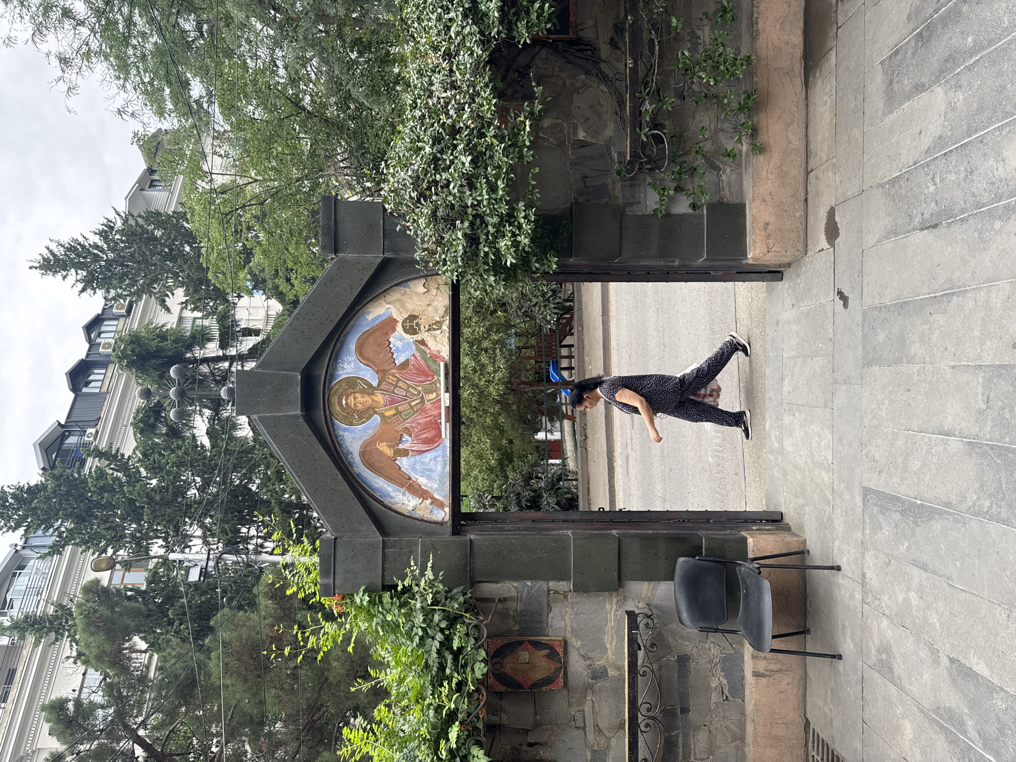 This is a vertical, eye-level shot of the entrance to a building with religious iconography in Tbilisi, Georgia. The foreground features a gray, stone tiled pathway leading to a dark stone archway. To the left of the archway, a black plastic chair sits on a raised stone platform. The wall to the left of the archway is made of dark stone and features a small, framed religious icon of a person in red and yellow robes.
The archway itself is dark stone with an arched fresco above the opening. The fresco depicts an angel with brown wings, wearing a red and pink robe, with gold trim and a halo. The angel is facing forward and holding a scroll. The opening of the archway reveals a street with vehicles and people in the background.
A woman in a black jumpsuit with white polka dots is walking through the archway from left to right. She is carrying a white bag in her right hand. Behind the archway, trees and a building with white walls and multiple windows are visible under a cloudy sky. Green foliage frames the archway on both sides.