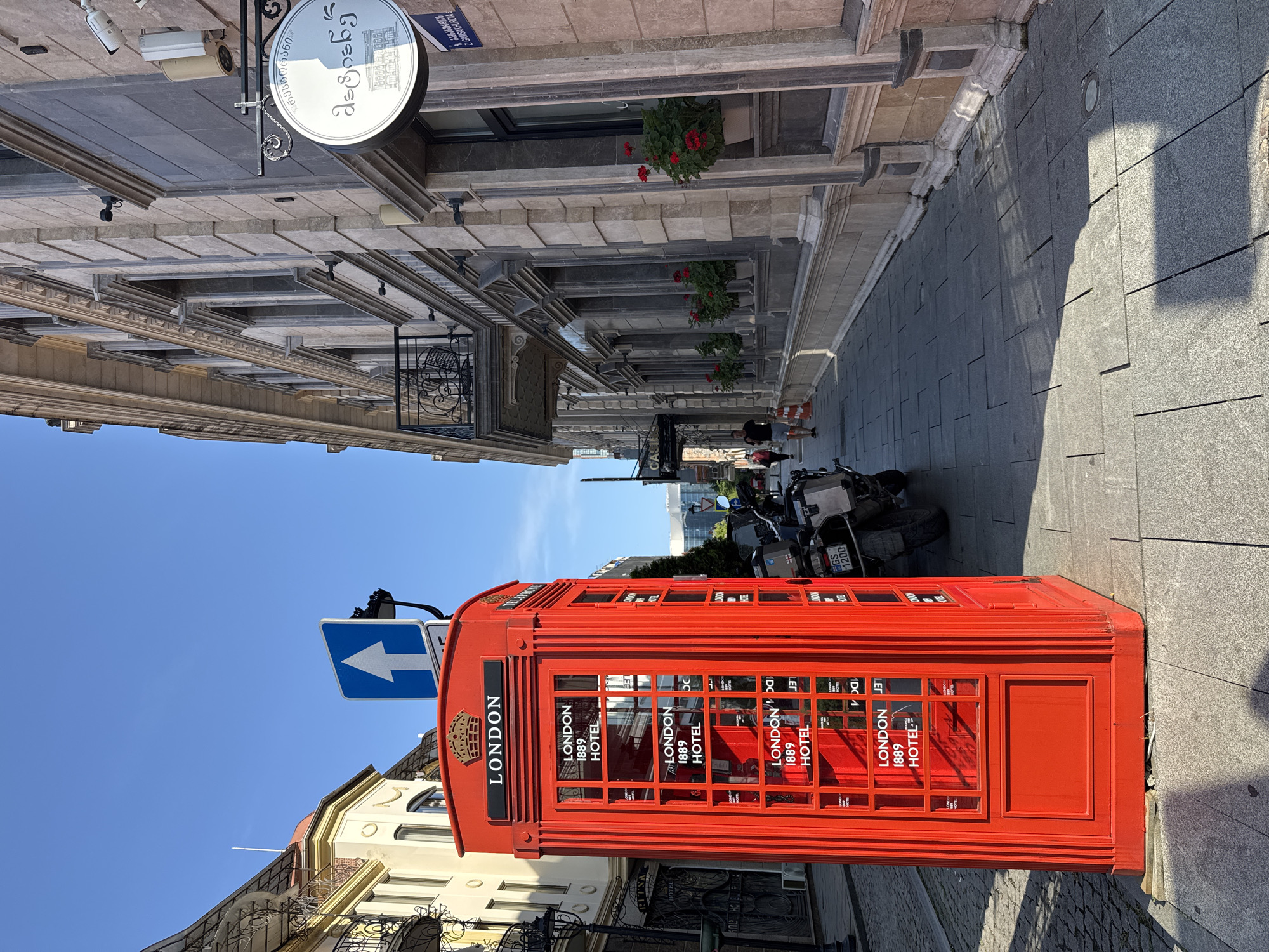 The image shows a red, traditional British telephone box on a city street in Batumi, Georgia. The telephone box is branded "LONDON 1889 HOTEL". To the right of the phone box is a building with a light-colored stone facade, featuring several windows and a balcony. A circular sign with Georgian script and a building illustration hangs from the facade. A street sign in Georgian script is also visible on the building. The street is paved with stone tiles, and a parked motorcycle is present next to the telephone box. In the background, a person is walking down the sidewalk. The sky is clear and blue.