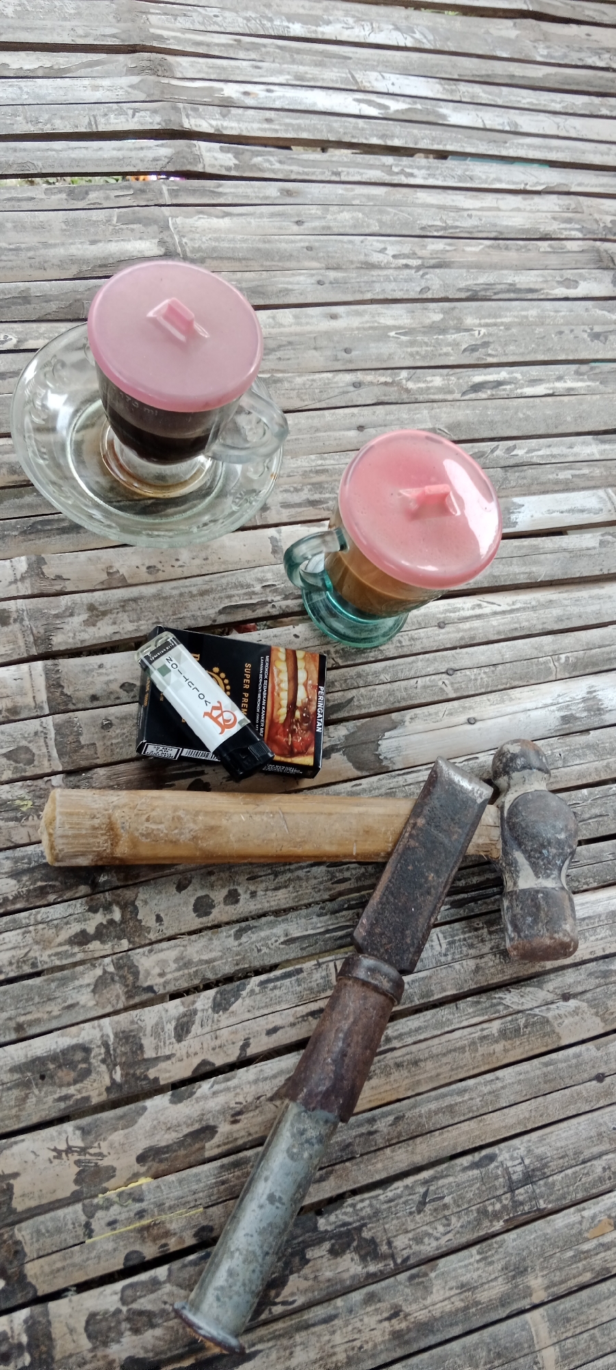 The image depicts a collection of items on a bamboo surface. Two glass cups, each with a pink lid, contain dark liquid, presumably coffee. Beside the cups is a pack of cigarettes and a lighter. A piece of bamboo, a metal chisel, and a small hammer are also present on the surface. The overall setting suggests an outdoor environment, potentially in Pelang, Indonesia, given the presence of bamboo. The scene appears to be staged, with the objects arranged for a photograph rather than captured in a candid moment. There are no people visible in the image.