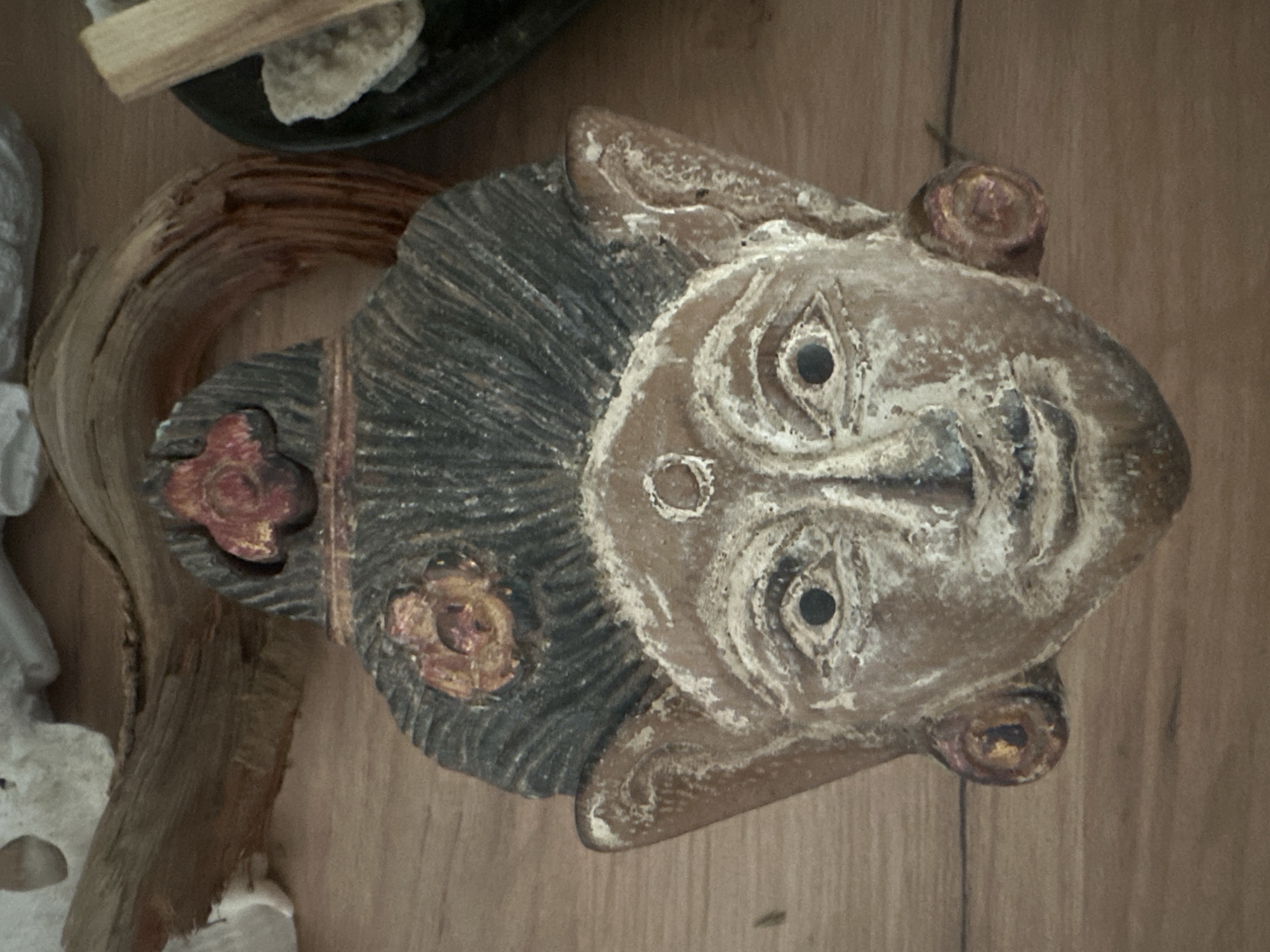 A carved wooden mask, likely of a spiritual or ceremonial nature, is placed on a wooden surface. The mask has a weathered, aged appearance, with dark carved hair, a prominent forehead with a circular marking, and almond-shaped eyes with dark pupils. Red and gold painted accents are visible on the mask, including decorative elements on the forehead and ears. To the left of the mask, a piece of gnarled, brown wood is positioned, with some white objects partially visible behind it. To the right, a dark bowl contains light-colored stones and what appear to be dried wood sticks, possibly palo santo. The overall setting suggests a decorative or ritualistic arrangement. The location of Novosyanovo, Russia is not depicted in the image.
