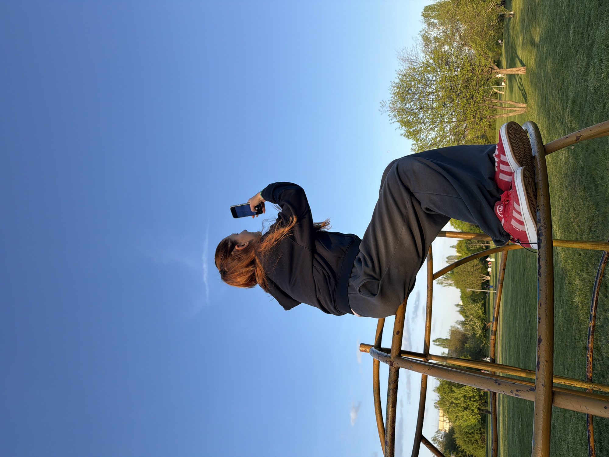 The media file displays a sideways view of a person sitting on a metal playground structure under a bright blue sky. The individual has long, reddish-brown hair and is dressed casually in a black hooded sweatshirt and dark, loose-fitting pants. They are holding a dark smartphone with both hands, pointing it upwards and away from their body, as if capturing a photo or video. Their feet, wearing red Adidas Campus sneakers with white stripes, are resting on a bar of the rusty, yellow-painted metal structure.
The sky, occupying the upper left portion of the image, is a clear blue with a faint wispy cloud trail. The lower right section reveals a park environment, characterized by expansive green grass and several trees, many showing signs of new spring growth. The lighting is soft and warm, indicative of late afternoon or early evening. The overall scene suggests a relaxed moment outdoors in a public park, consistent with a location such as Sofia, Bulgaria. The only visible text is the "CAMPUS" label and Adidas logo on the sneakers.