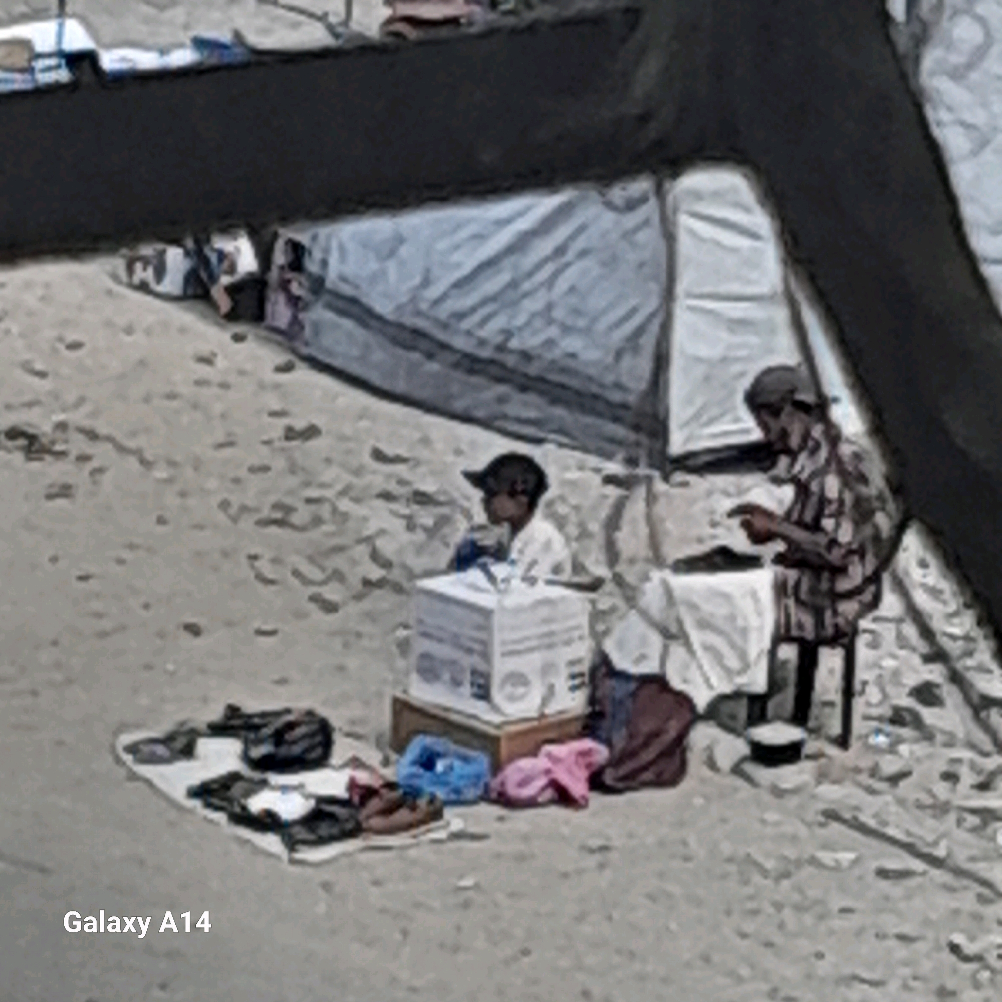 In Khan Yunis, Palestinian Territories, a scene unfolds on sandy ground amidst makeshift shelters. In the foreground, scattered items of clothing and bags lie on a light-colored fabric. To the left, a young child wearing a dark cap and a light-colored shirt sits near a white cardboard box. To the right, an adult wearing a cap and a patterned shirt sits on a dark stool, appearing to be engaged in an activity with their hands, possibly examining an object. In the background, tents are visible, suggesting a temporary settlement or displacement camp. The overall environment is one of arid terrain with scattered debris.