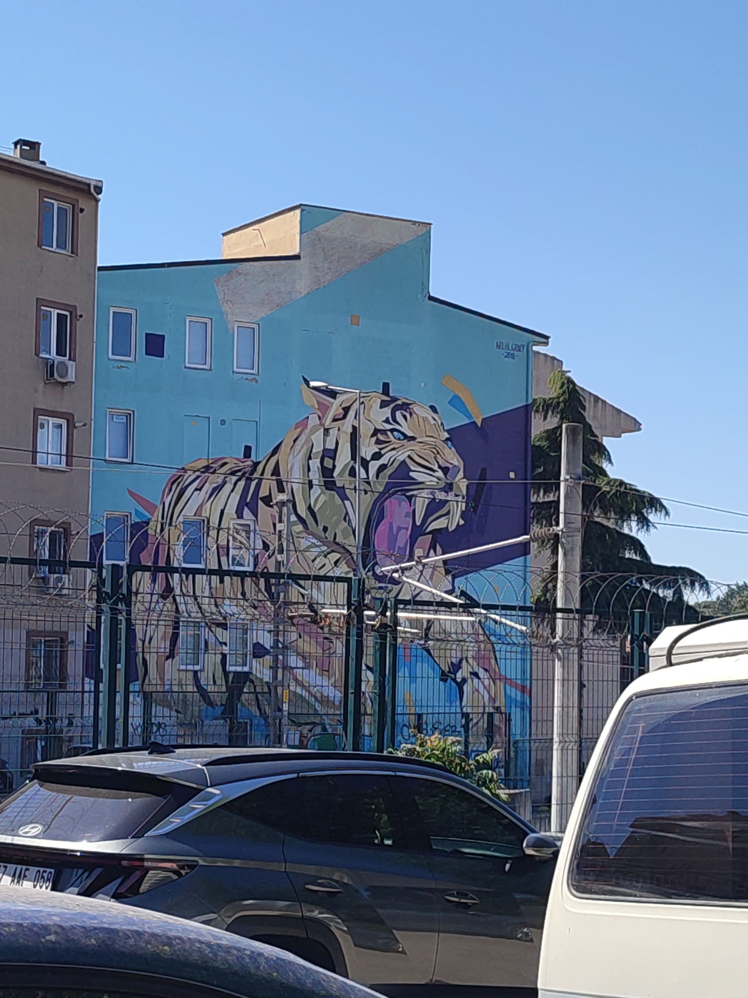 A large mural of a tiger in a stylized, geometric design adorns the side of a light blue building. The tiger's mouth is open, revealing fangs and a purple tongue, and its eyes are blue. The mural is visible behind a metal fence with barbed wire. To the left of the blue building is a light brown building with several windows and an air conditioning unit. In the foreground, a dark gray SUV is parked, partially obscuring the fence and mural. The sky is clear and blue. The mural is located on the side of a residential building in Kadıköy, Turkey.