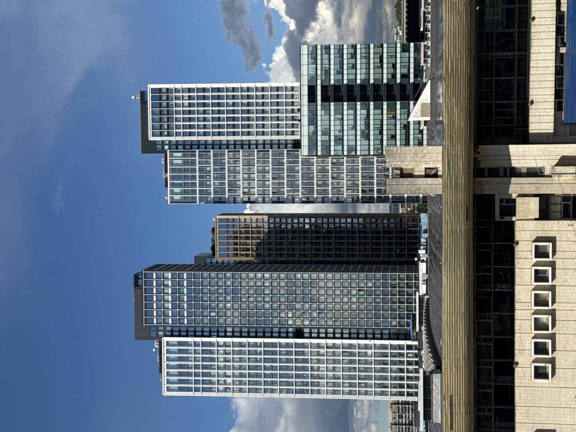 The image shows a cluster of modern high-rise buildings against a backdrop of a blue sky with scattered clouds. In the foreground, the roof of a lower, older building with a textured, horizontal design stretches across the lower third of the frame. This building has large, dark-tinted windows. Above this, the prominent skyscrapers vary in their facade design. The tallest building on the left is predominantly white with a grid of windows. Next to it, a dark glass skyscraper has a repeating grid pattern of square windows. To the right of that, a narrower building features a warm, brownish-toned facade with a similar window pattern. The rightmost tall building is a combination of white, grid-patterned sections and a lower, wider section with dark glass and horizontal dividing lines. A smaller, glass-enclosed building is visible at the base of the rightmost skyscraper. The overall scene depicts an urban landscape with a contrast between older and newer architectural styles. The location is identified as Khimki, Russia.