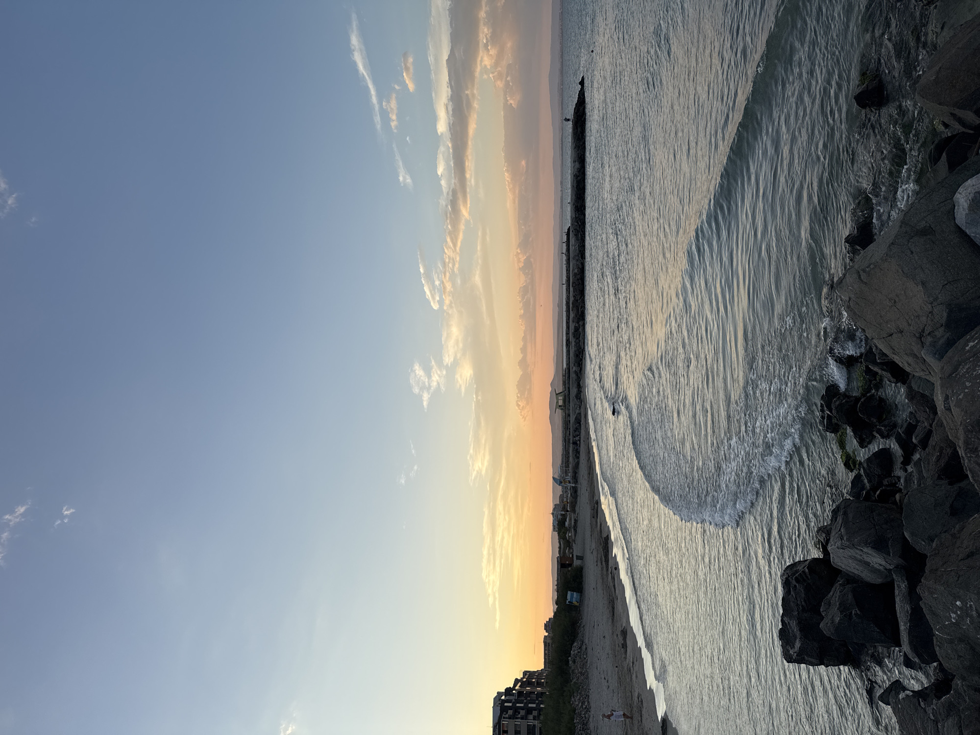 The image depicts a sunset over the Black Sea coastline in Pomorie, Bulgaria. In the foreground, dark, wet rocks are visible, with the sea gently lapping against them. A wave is breaking closer to the shore. The sandy beach stretches to the left, where a few dark structures, likely buildings, are situated. A person in white clothing stands on the beach.
Further out, a breakwater extends into the sea, with several small figures standing on it, possibly fishing or enjoying the view. The water transitions to a calmer, deeper blue as it meets the horizon.
The sky is a gradient of colors, transitioning from a soft orange and yellow near the horizon, indicating the setting sun, to a clear, pale blue overhead. wispy, light-colored clouds are scattered across the upper portion of the sky. The overall scene conveys a peaceful and serene atmosphere.