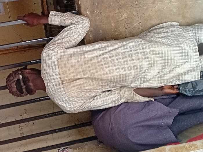 The image, taken in Ayama, Nigeria, depicts two individuals at a barred opening. In the foreground, a male subject is viewed from the rear, wearing a light-colored, faintly plaid long-sleeved tunic. A reddish-brown, textured cap is worn backward on his head, exposing dark hair beneath. His left hand grips a vertical metal bar, part of a series forming a barrier. His right hand holds a black plastic bag positioned at his side. A faint discoloration is visible on the upper back of his tunic. Partially obscured to his left and slightly behind him, a second individual is visible, dressed in dark blue or purple patterned attire, appearing to bend forward. The background consists of a light brown, worn wall. Faded white lettering is discernible on the left side of the wall and the bars. To the right of the main subject's torso, distinct white graffiti reads "17/2/2020 Labiyaffa". The area beyond the vertical bars appears dark.