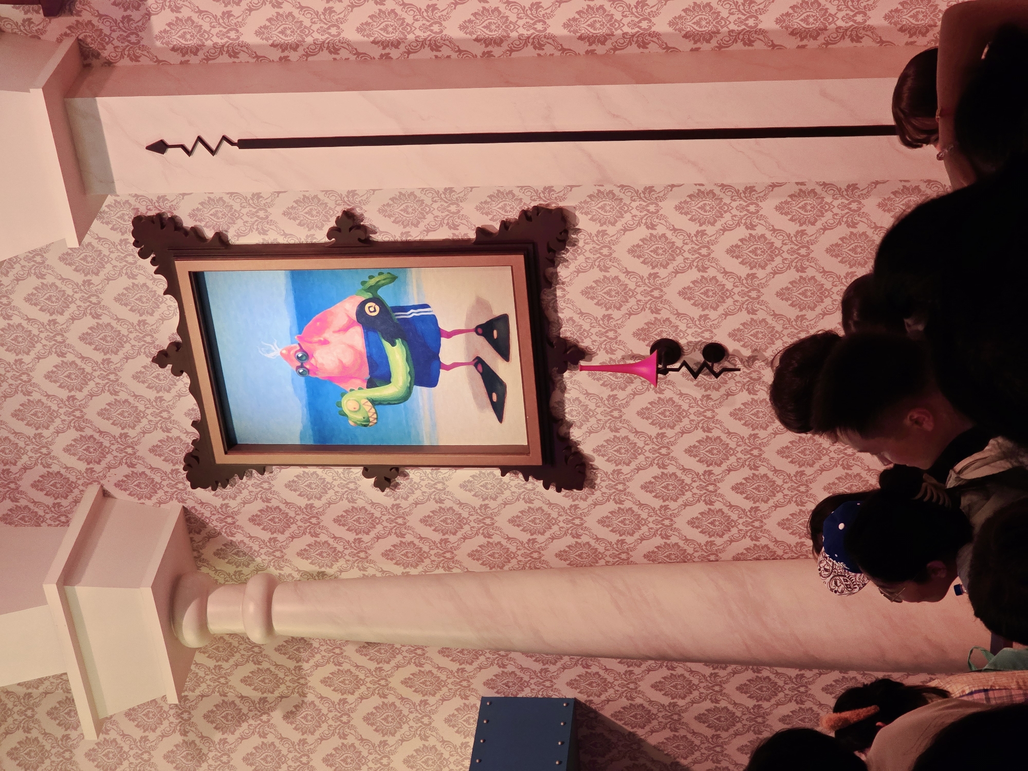 The image shows a dimly lit room with patterned wallpaper. A large framed painting hangs on the wall, depicting a pink, anthropomorphic character wearing sunglasses and swim trunks, holding a green inflatable dinosaur. To the left of the painting is a tall, decorative column. To the right is another column with a black, zig-zagging decorative element. In the foreground, several people are visible from behind, their faces mostly obscured, suggesting they are observing the artwork or the room. The overall impression is that of an indoor exhibition or themed space. The image is believed to be from Tongzhou District, China.