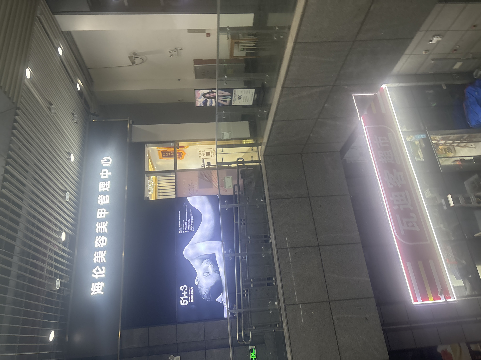 The image is a medium-angle shot of an indoor commercial space, possibly a shopping mall or a multi-level retail area, located in Liwan District, China. In the upper section, a large black sign displays white Chinese characters that read "海伦美容美甲管理中心" (Hailun Beauty Nail Management Center). Below this sign, a large digital advertisement features an image of a person's upper body lying down, with text in both Chinese and English, including "51+3 HYALU COMPLEX™" and promotional slogans. A metal railing with decorative patterns is visible in front of this advertisement. To the right, a vertical poster displays an image of a woman and the number "999". Further to the right, there is a white wall with some fixtures and a small wooden table with stools. In the lower section of the image, a bright, illuminated red sign with white Chinese characters, "瓦迪客超市" (Wadike Supermarket), is prominently displayed. The environment appears to be dimly lit, with the neon lights of the signs providing the primary illumination. The architecture includes textured walls, a suspended ceiling with horizontal panels and circular lights, and glass railings. There are no people clearly visible in the frame.
