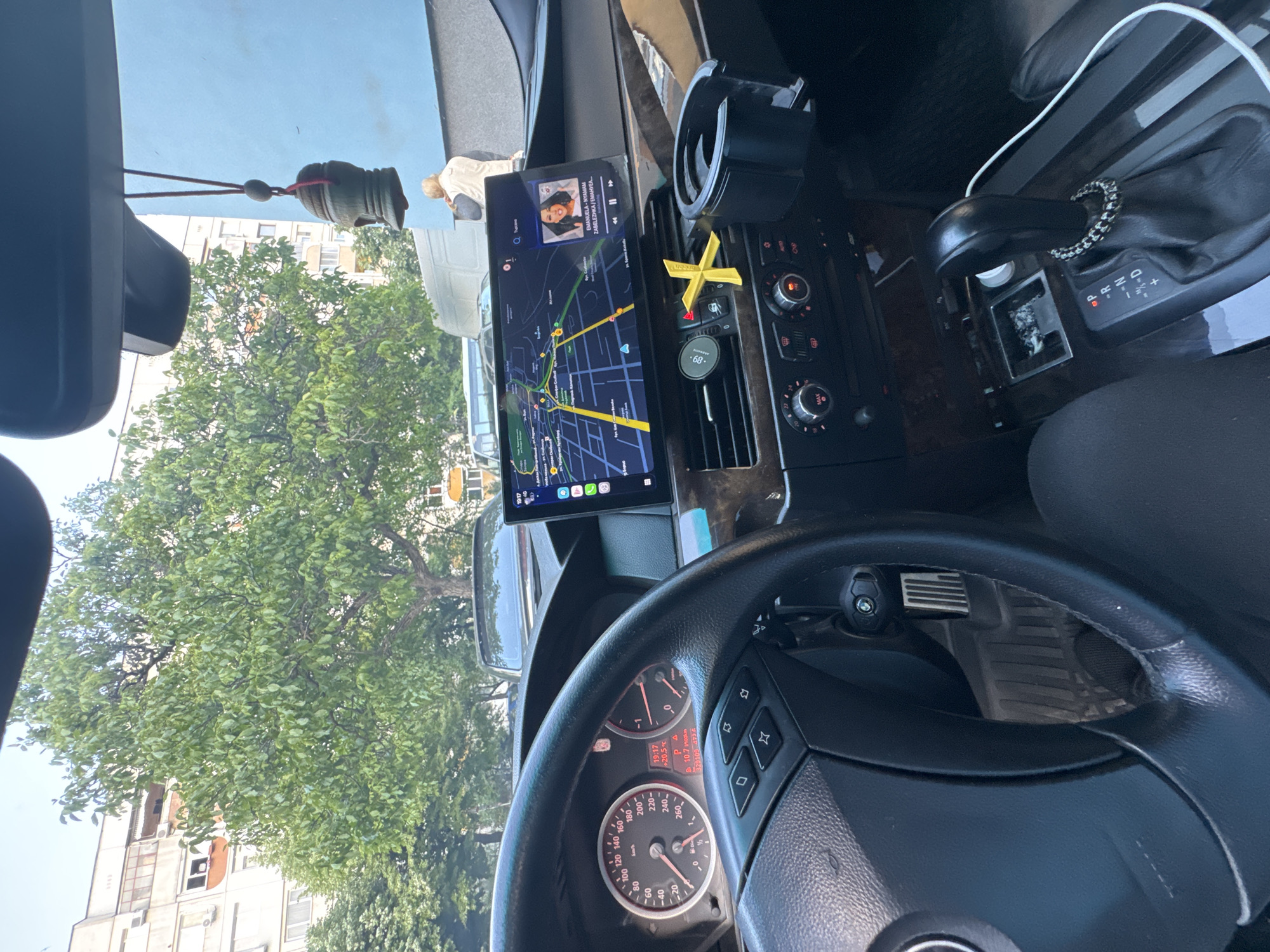 The image is taken from inside a car, showing the dashboard, steering wheel, and a view through the windshield. The car appears to be parked or stopped, with a large tree and buildings visible outside. A navigation app is displayed on a screen mounted on the dashboard, indicating a location within the city of Stara Zagora, Bulgaria. The app is playing a song by Emanuela-Nyamam called "Zabelezhka/Eмануел". The car has a manual transmission and a digital speedometer displaying 10.7 km/100km. There is an air freshener in the shape of an "X" attached to the air vent.