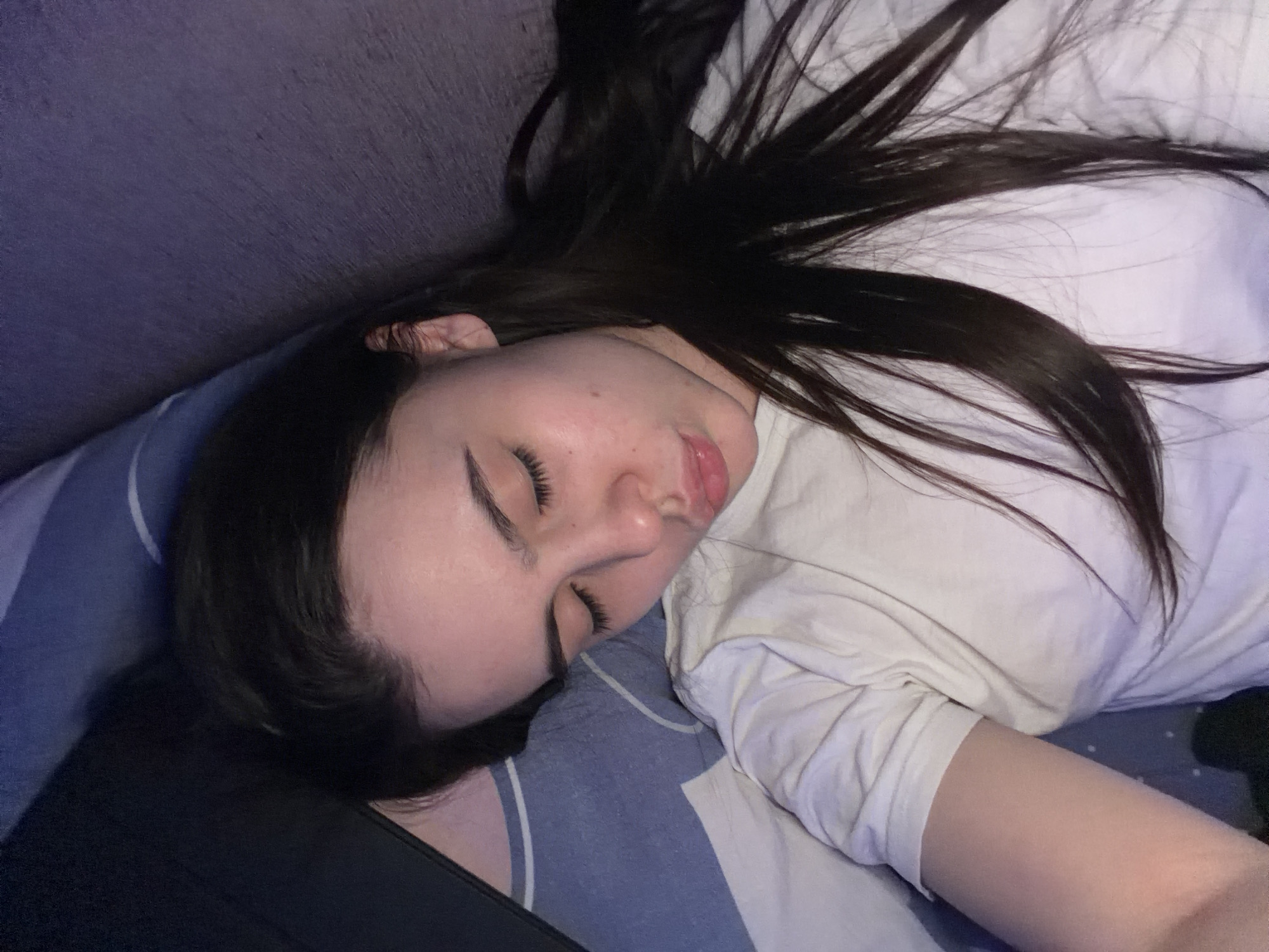 A woman is lying down, presumably asleep, with her head resting on a blue pillow with white stripes. Her long, dark hair is spread out around her head and onto a white t-shirt she is wearing. The background includes a dark grey or purple surface, possibly a sofa or wall. A black rectangular object is also partially visible in the lower-left corner. The location cannot be determined from the visual information provided. There is no indication that the scene is in Chaikovsky, Russia.