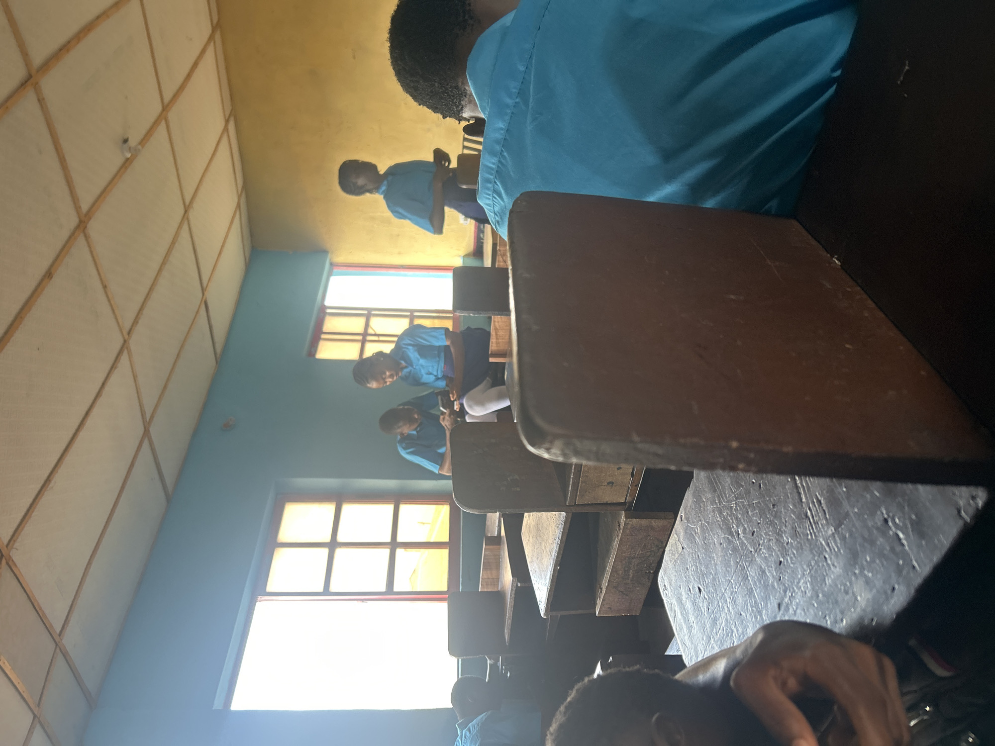 The image depicts an indoor scene within a classroom in Ikorodu, Nigeria, shot from a low perspective, looking towards the front or side of the room. The photograph is oriented sideways, but when rotated, it shows several students, primarily young people, dressed in light blue uniforms, seated at wooden desks and benches.
In the immediate foreground, part of a student's head and hand are visible, seated at a dark, worn wooden desk. Beyond this, rows of similar wooden desks and attached benches extend into the room. Bright sunlight streams intensely through large, multi-paned windows on the left side of the frame, creating significant glare. The walls of the classroom are painted in two distinct colors: a light blue where the windows are situated, and a warm yellow on the opposite wall. The ceiling features a grid pattern, possibly constructed from light-colored panels with wooden supports, and a single light bulb is faintly visible.
Further back in the classroom, one student is seen standing or leaning near their desk, appearing to hold a small object, perhaps a phone or book. Other students are seated in various positions, some looking forward. The overall activity suggests a routine school day, with students either attending a lesson, studying, or waiting. The strong sunlight indicates it is daytime. The setting appears modest but functional, typical of a local school environment. No discernible text is visible in the image.