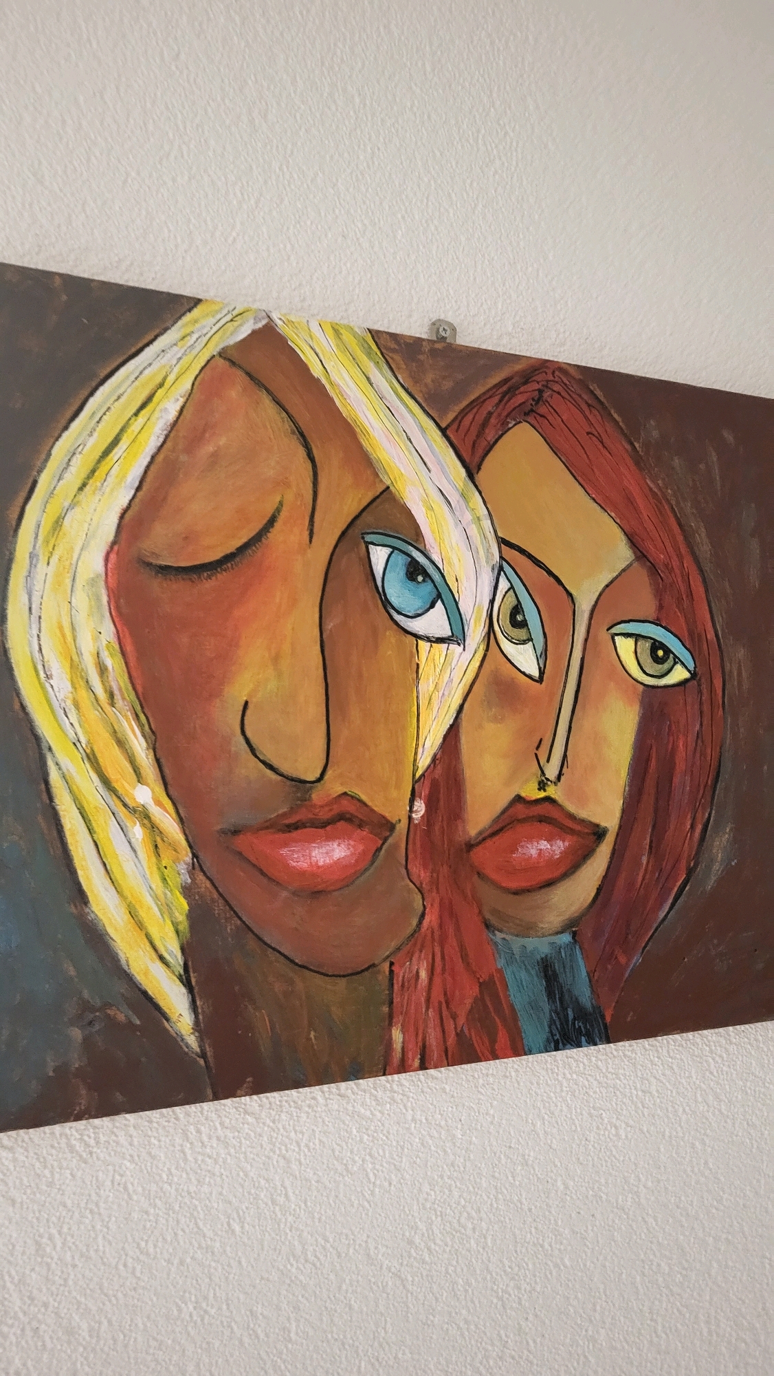 A horizontal painting hangs on a textured white wall. The painting is an abstract portrait of two women. The woman on the left has blonde hair, closed eyes, and a tear falling from her eye. The woman on the right has reddish-brown hair and her eyes are open, with one eye facing forward and the other looking slightly to the side. Both women have red lips and dark outlines around their faces. The background is a mottled brown. A small silver screw is visible in the top center of the painting, attaching it to the wall. The wall appears to be an interior wall, possibly in a home or gallery. The scene is devoid of any indication of the city of Općina Tučepi, Croatia.