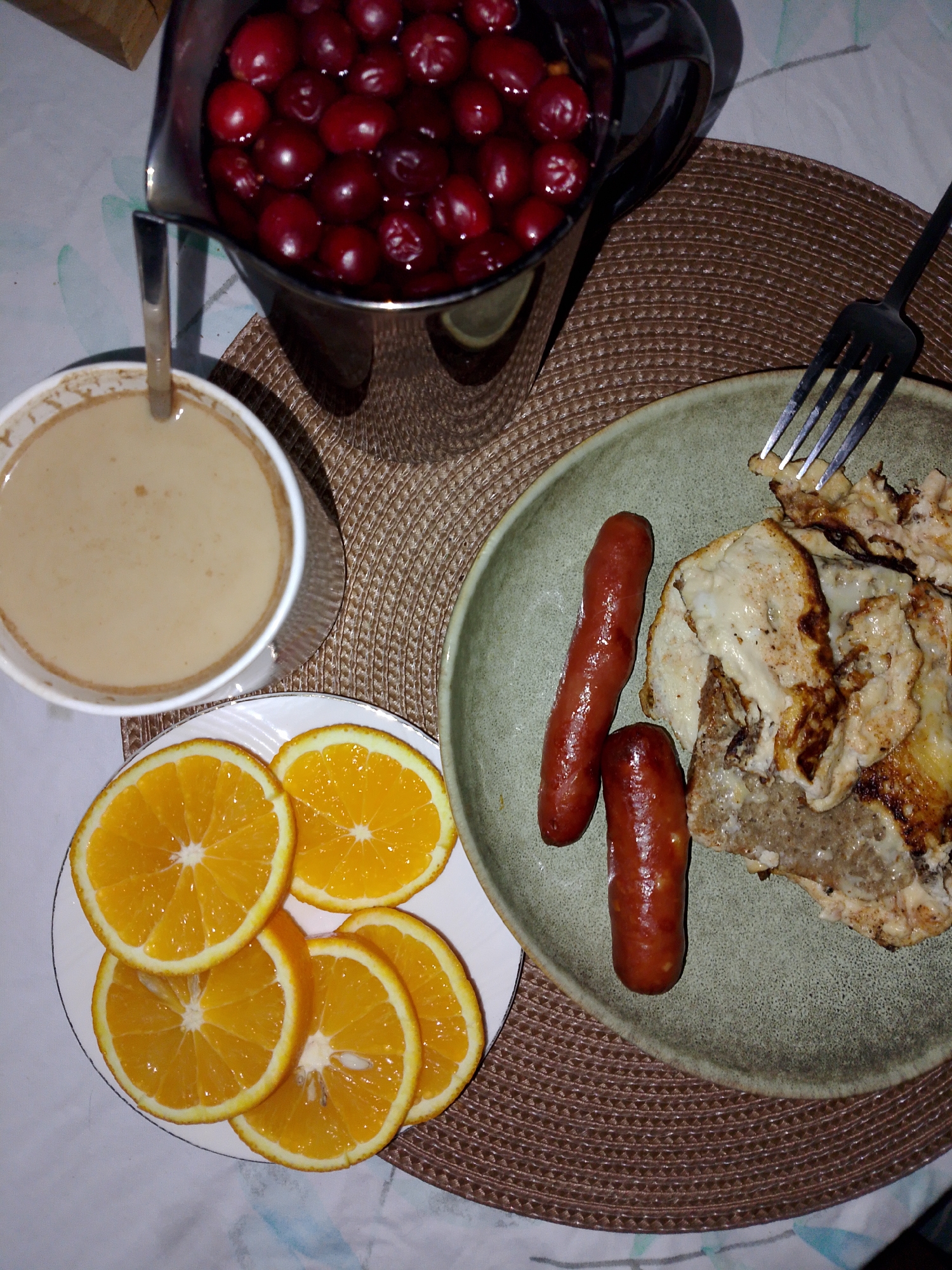 On a patterned placemat, a breakfast setting is arranged. A stainless steel pitcher filled with dark red cherries sits near a white mug containing a beige liquid and a spoon. A white plate holds several orange slices. A green plate includes two sausages and scrambled eggs, with a fork placed near the eggs. The setting appears to be indoors, potentially on a table. The location is not discernible.