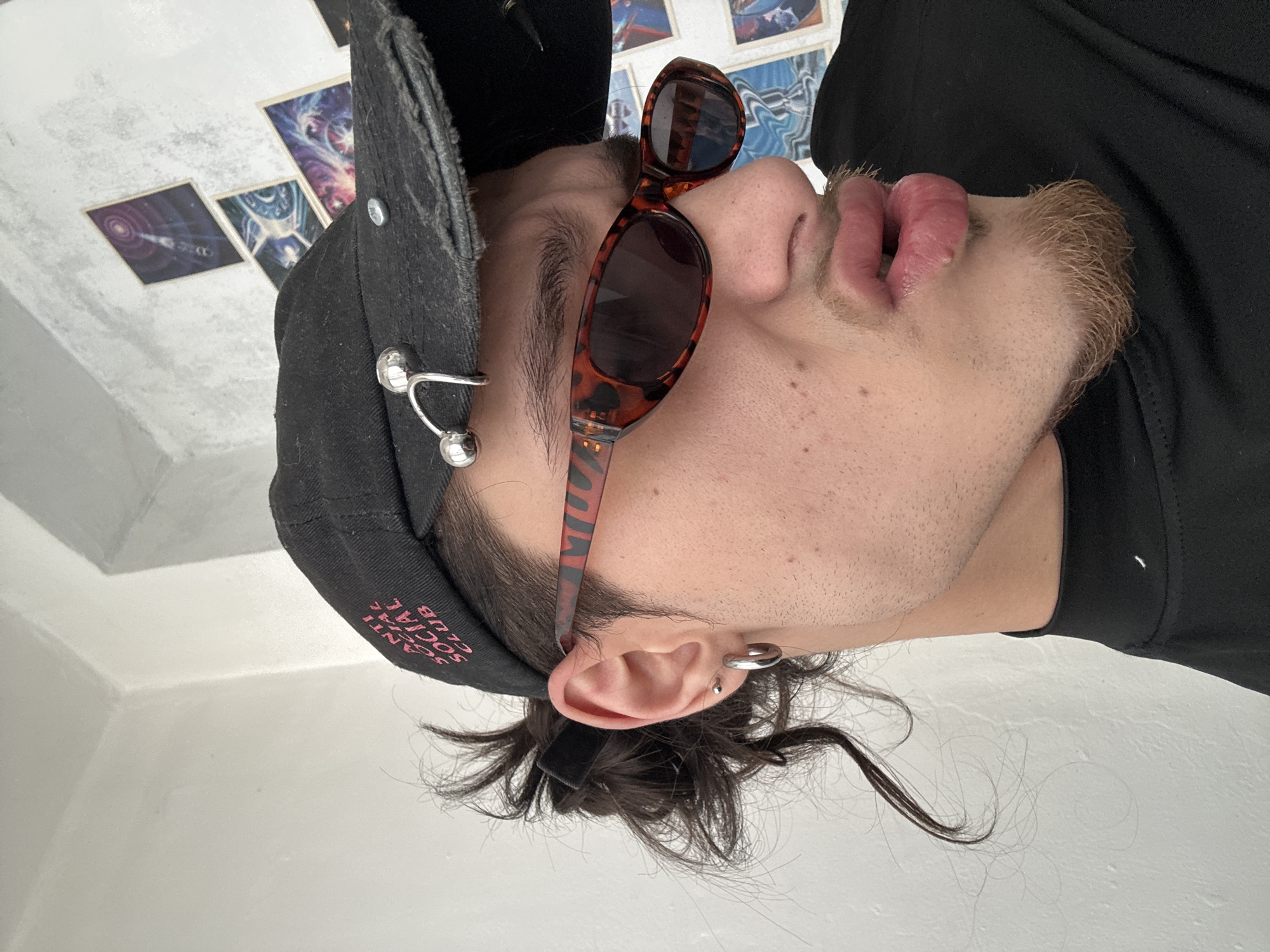 A low-angle portrait captures a person in profile. The individual wears a black baseball cap adorned with silver ball hardware, tortoiseshell sunglasses resting on the nose, a black t-shirt, and has ear piercings. The face is visible, showing stubble and freckles. In the background, there are posters on a textured white wall. The location is unknown and not discernible from the image.