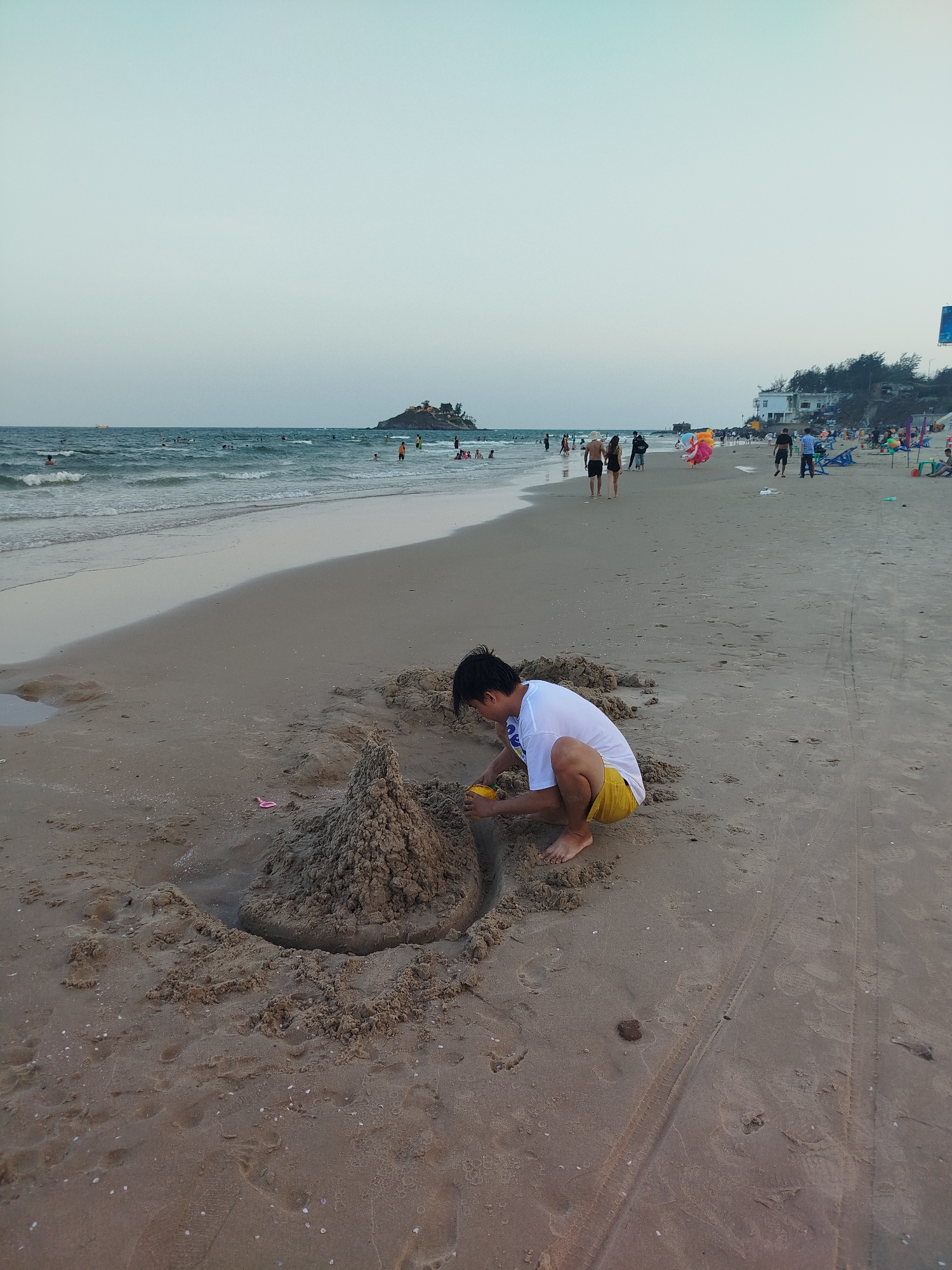 A young person is building a sandcastle on a beach. The person is wearing a white t-shirt and yellow shorts. They are squatting near a sandcastle, using a small yellow tool to mold the sand. The sandcastle is in the foreground. The beach stretches out in the background, with the ocean on the left. People are walking and swimming in the distance. A building is visible on the right side of the image. The sky is a pale blue. The image was taken in Ho Chi Minh City, Vietnam.