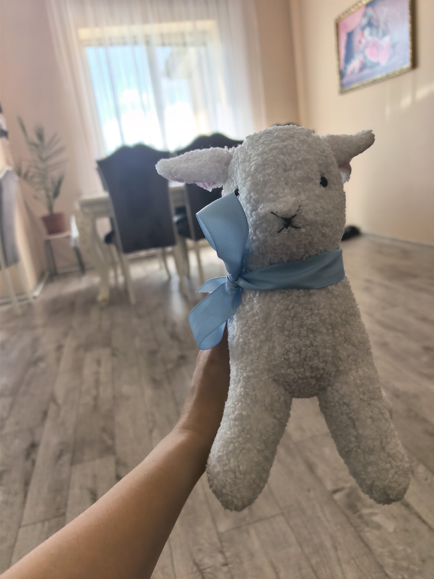 A fluffy white lamb stuffed animal with a blue ribbon around its neck is held up by a person's arm. The lamb has black embroidered eyes and nose. In the background, a dining table with chairs is visible, along with a window with sheer curtains and a framed picture on the wall. The floor is made of light-colored wooden planks. The scene appears to be indoors, possibly a dining room or living area.
