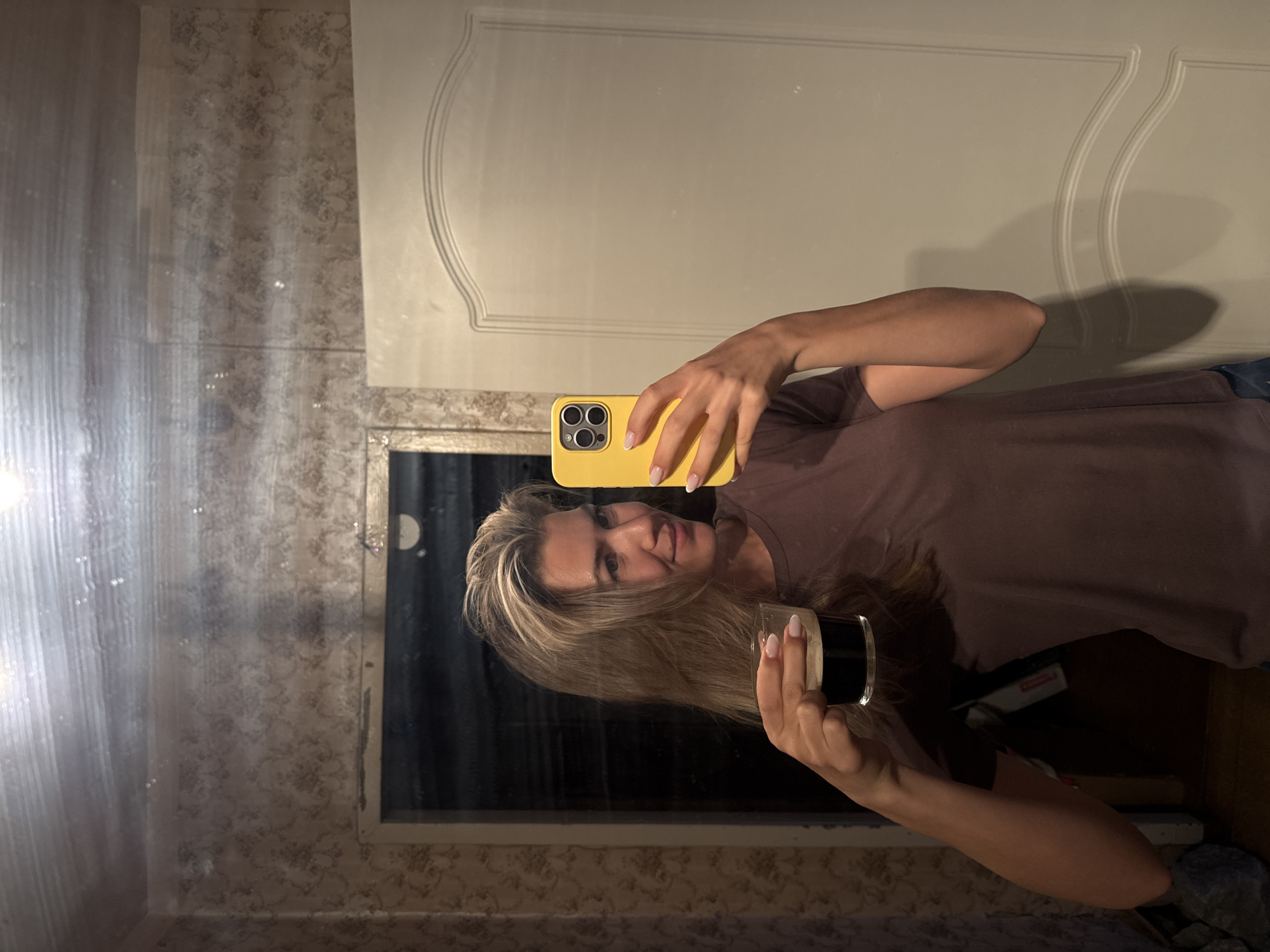 A woman is captured in a mirror selfie, holding a bright yellow smartphone in her right hand and a glass with a dark beverage topped with white foam in her left hand. She has long, straight blonde hair, fair skin, and is wearing a plain brown t-shirt. Her nails are manicured with a light, possibly French tip, polish. She is smiling faintly at her reflection.
The background visible in the mirror behind her reflection shows a room with patterned wallpaper, possibly a floral or damask design, on the left. To the right, a white door with decorative raised paneling is visible. The reflection also shows what appears to be a dark, rectangular object below the mirror, possibly a window or a dark surface, and a light-colored counter or shelf beneath it. There's a subtle bright reflection on the far left side of the mirror, possibly from an external light source or window. The overall lighting is somewhat dim, suggesting an indoor setting, potentially in the evening or with artificial light. No discernable text is visible.