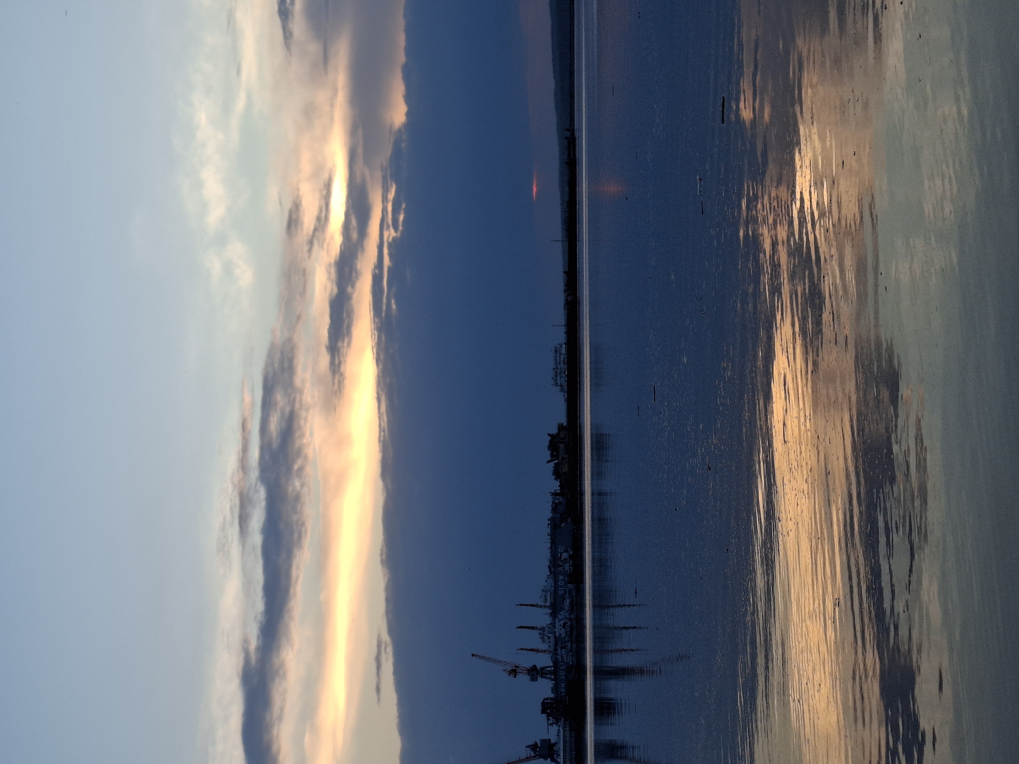 The image depicts a waterfront scene in Chaikovsky, Russia, at dusk or dawn. Cranes and industrial buildings are visible along the shoreline. The sky is a gradient of light blue to dark blue with clouds. Reflections of the sky and the city are visible in the water.