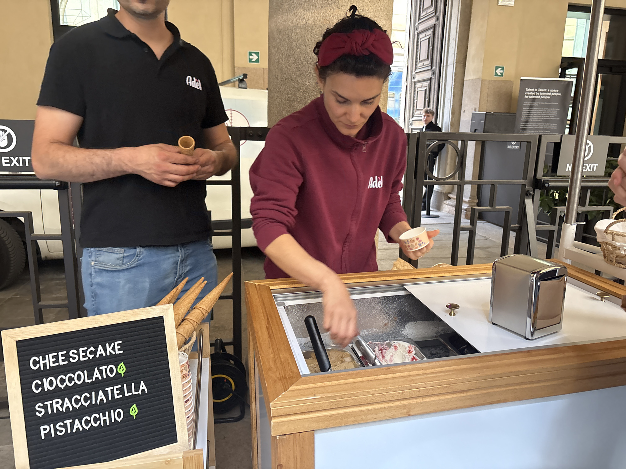 The scene is an outdoor ice cream stand operated by "Adel" in Milan, Italy, offering several gelato flavors. A woman in a maroon jacket and matching headband is scooping gelato into a small paper cup from an ice cream freezer. To the left, a man wearing a black polo shirt with the "Adel" logo holds an empty cone. A menu board lists the flavors: Cheesecake, Cioccolato (Chocolate), Stracciatella, and Pistacchio, each marked with a leaf symbol. Cones are displayed next to the sign. Behind the stand are barriers with exit signs, leading into a building. In the background, a sign reads "Talent to Talent: a space created by talented people, for talented people." A man is visible exiting the building in the background.
