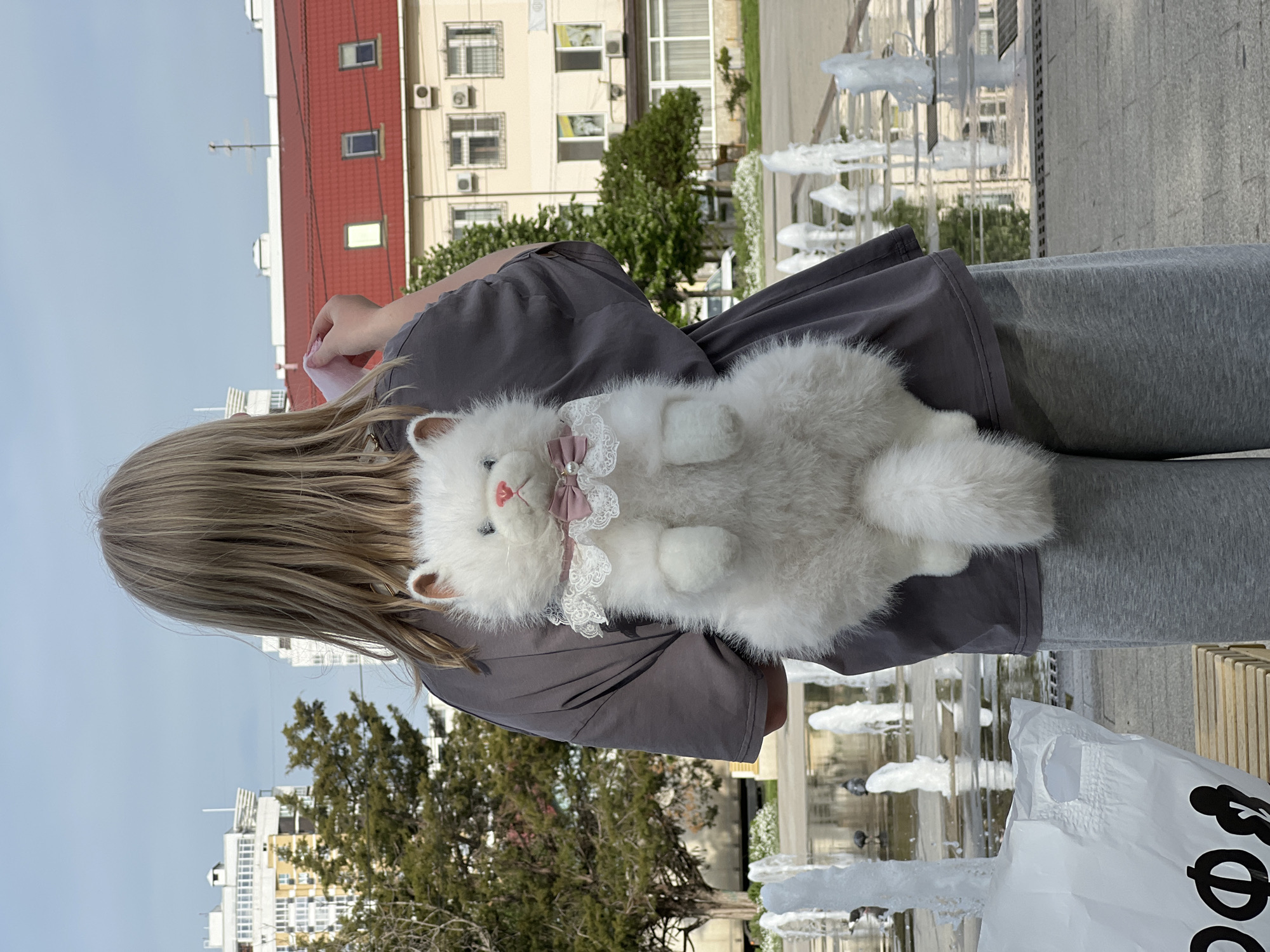 A girl with shoulder-length blonde hair is standing outside in Krasnodar, Russia. She is wearing a dark gray t-shirt and light gray sweatpants. She is holding a fluffy white cat plush toy with a white lace collar and a small pink bow. Behind her is a fountain, and in the background are buildings with red roofs and trees. There is also a white plastic bag on the right.