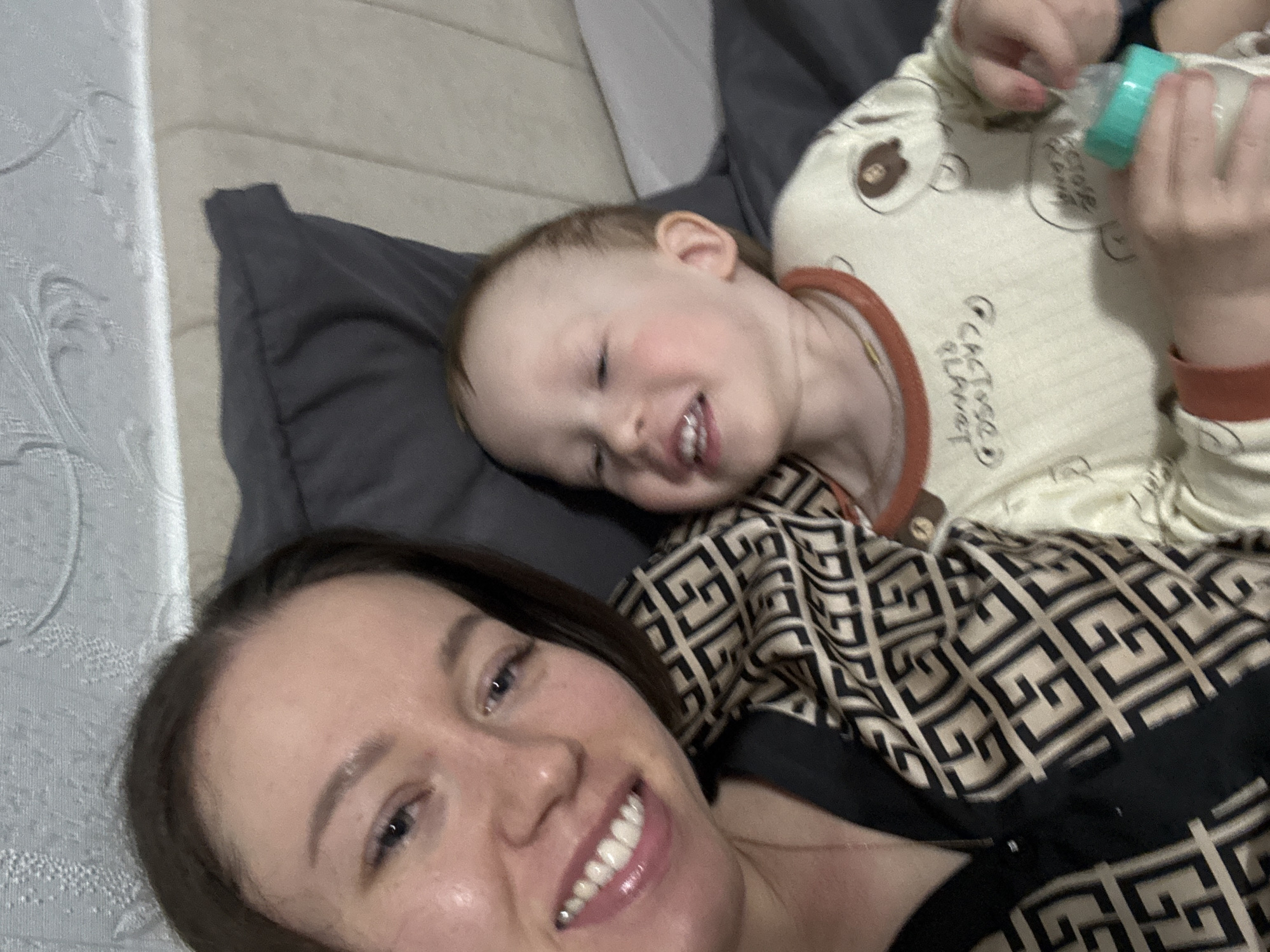 A selfie shows a woman with dark hair and a fair-skinned child lying on a gray pillow. The child is smiling and holding a baby bottle. The woman is also smiling. The scene appears to be indoors, likely in a domestic setting. There is light gray bedding visible in the background. There is no information to suggest the image was taken in Ferma, Russia.