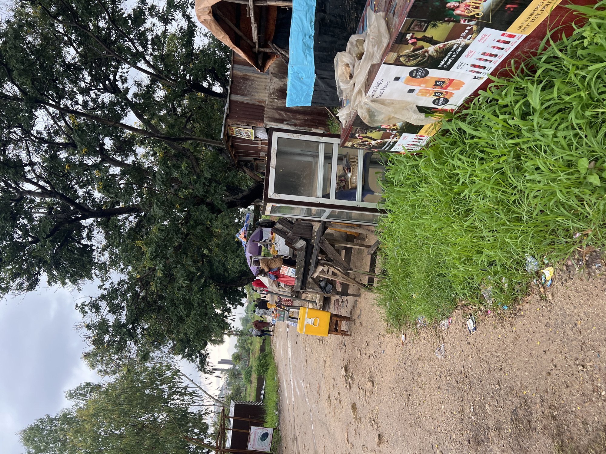 This image depicts an outdoor scene, likely a roadside market or informal settlement in Rafin Par, Nigeria. The foreground is dominated by a dirt path with scattered debris and a patch of lush green grass. To the right, a large display board advertising Coca-Cola products and other beverages is propped up. The board features vibrant images of bottles and cups, along with prices in Nigerian Naira (N).
Further into the scene, a makeshift stall is visible, constructed from corrugated iron and wood. Various items are displayed on tables, including what appear to be packaged goods and possibly food items. A prominent yellow cooler box sits on a stool in front of the stall. Several people are gathered around the stall, engaged in what seems to be commercial activity, though their faces are not clearly discernible. Some are standing, while others are seated. The atmosphere appears casual and busy, characteristic of a market day.
Behind the stall, a large, leafy tree provides shade. Further back, more trees and some distant buildings can be seen, suggesting a semi-urban or rural fringe setting. The sky is overcast, indicating a cloudy or possibly rainy day. The overall impression is one of everyday commerce and community life in a developing area.