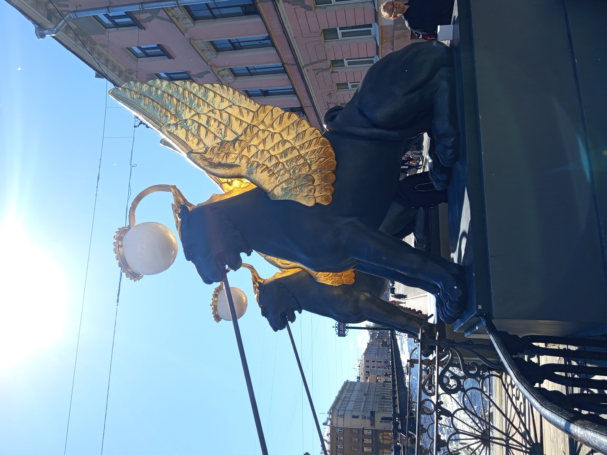 The image shows a low-angle, outdoor view of two griffin statues, their dark bodies contrasted with gleaming golden wings. One griffin is more prominently featured, its head and upper body visible, facing to the left. The other griffin is partially visible behind it. Both statues appear to be holding lamps with large, white, spherical globes. They are positioned near a decorative black metal railing.
In the background, the blue sky is clear, with the bright sun causing lens flare in the lower left corner. Above the statues, part of a building with a pink facade and dark-framed windows is visible, suggesting an urban setting. Further in the background, other buildings and a body of water can be seen, possibly a canal or river.
A few people are visible in the distance, appearing as small figures on a walkway or street. One person, a woman wearing glasses and a black top, is closer to the foreground, looking to the left.
The scene is illuminated by bright sunlight, indicating it is daytime. The overall impression is one of grandeur and artistic detail, with the statues being the focal point. The location appears to be Saint Petersburg, Russia, given the architectural style of the buildings and the presence of canals. No visible text is present in the image.
