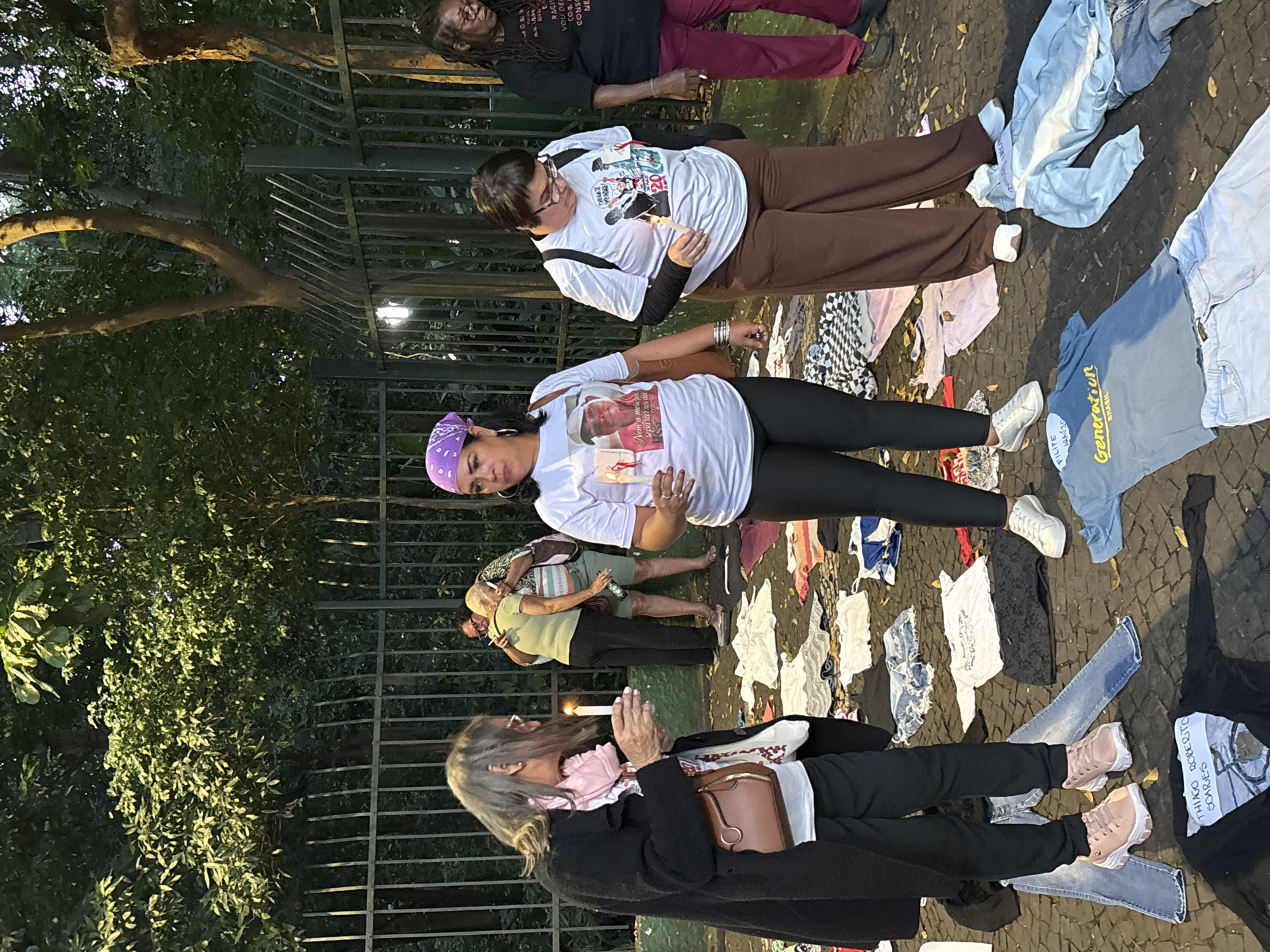 Several people are gathered in what appears to be a park or public space. They are standing amidst an array of clothing items laid out on the ground. Four individuals are distinctly visible. All the people are white except for one, a black woman. One woman is wearing a purple bandana, a white shirt with a photograph, black pants, and white tennis shoes, and she is holding a lit candle. Another person is wearing a white shirt with a graphic and brown pants. A third person is wearing a black overcoat and has a shoulder bag. The fourth individual is a black woman with burgundy pants and is wearing a black shirt with printed text. Some of the clothing items on the ground have writing or labels, including one with the words "Generation Brasil" and one with "THIAGO ROBERTO SOARES". The overall impression suggests a somber gathering or vigil.