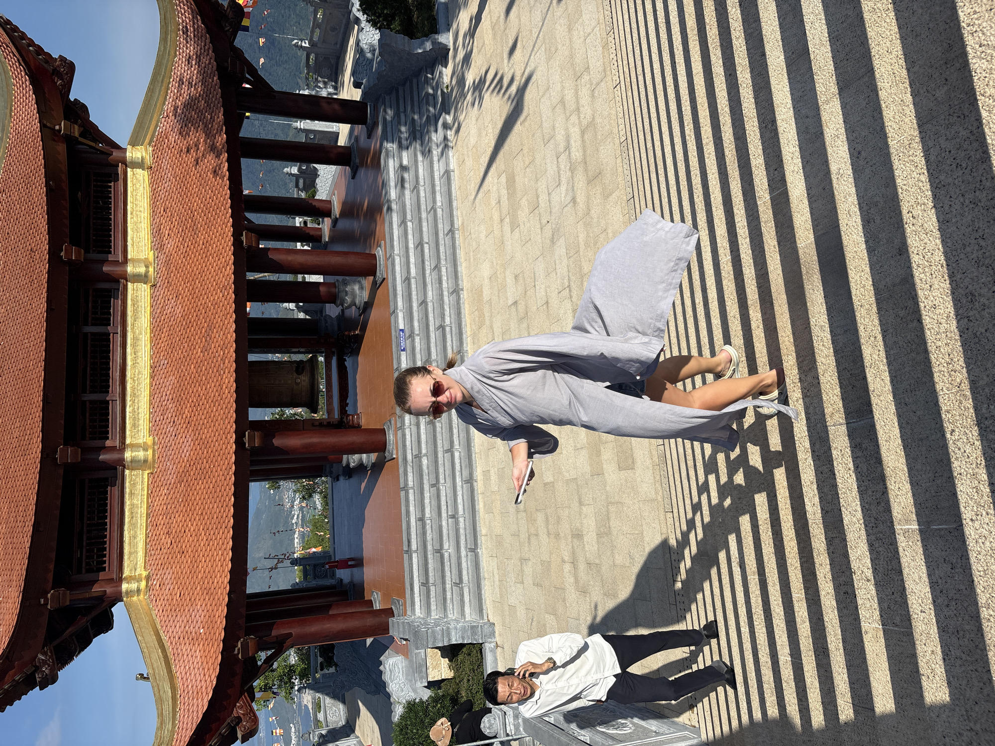 A woman in a grey robe and sunglasses holds a phone while walking on a paved surface. She is facing the camera. A man in a white shirt and black pants is lying on the same surface, talking on a cell phone. A traditional building with a red and gold roof is visible on the left. The environment suggests a location in or near the city of Diên Khánh, Vietnam, based on the architecture and landscape. Sunlight creates strong shadow patterns on the ground.