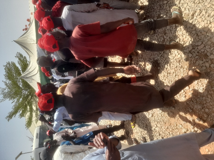The scene is an outdoor gathering in Gusau, Nigeria (latitude: 12.156, longitude: 6.709). The gathering is in a dusty, rocky area, with a few white tents in the background. Many people are present, with most wearing red caps, possibly signifying a group affiliation. Some people are wearing traditional clothing, while others are wearing more modern attire. The people are interacting and moving around, and the atmosphere seems casual. The image captures a snapshot of a social event or gathering in Gusau.
