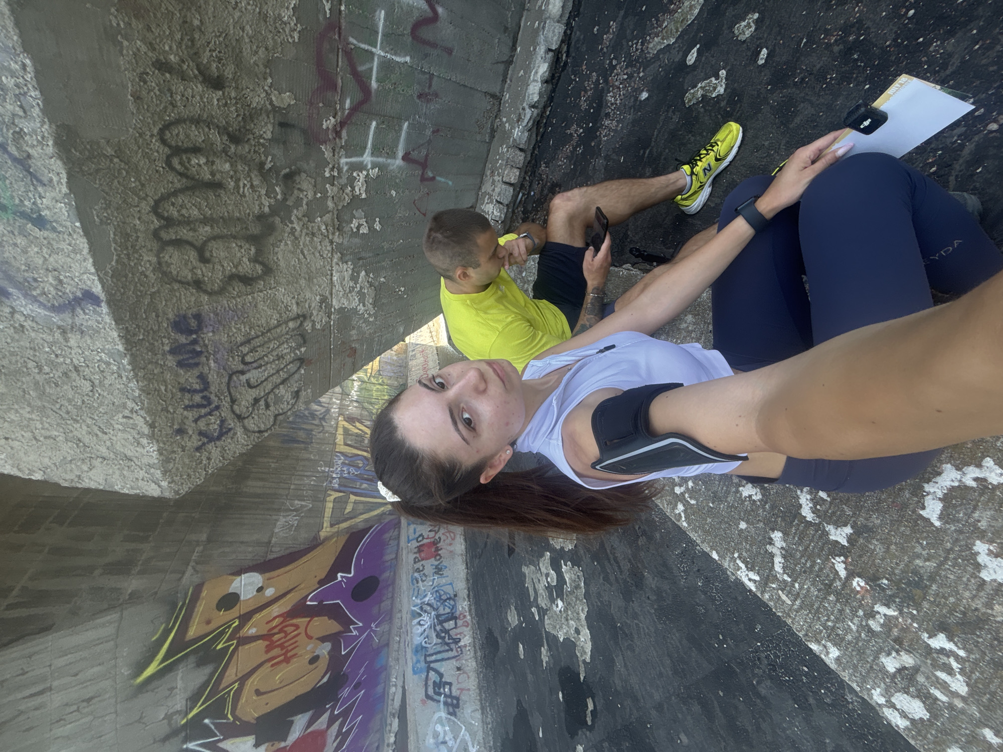 In Varna, Bulgaria, beneath a graffiti-covered concrete structure, a young woman with dark hair pulled into a ponytail, wearing a white athletic top and dark leggings, is in the foreground of the image, holding a phone arm band on her left arm. She is looking directly at the camera. To her right and slightly behind her, a man with short hair, wearing a neon yellow t-shirt and dark shorts, sits on the concrete, looking down at his phone. The environment is characterized by rough concrete walls adorned with vibrant graffiti, and a dark, textured ground surface. The overall impression is a candid outdoor shot, possibly during a break from physical activity.
