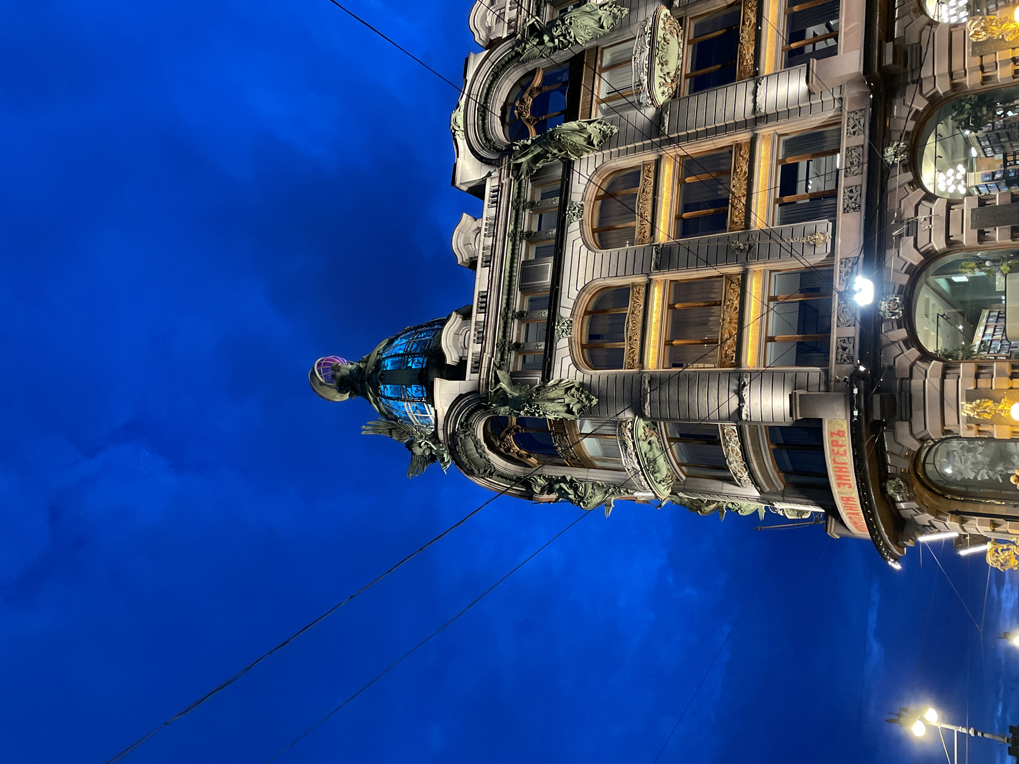 The image captures an architectural detail of the Singer House in Saint Petersburg, Russia, under a dark blue sky with visible clouds and power lines. The building features ornate Art Nouveau design, with a prominent domed roof structure illuminated by blue and purple lights. Decorative elements, including winged figures and intricate carvings, adorn the facade. The lower levels of the building display arched windows and the inscription "КОМПАНИЯ ЗИНГЕРЪ" in red lettering. Streetlights and interior lights of the building are visible, suggesting it is evening or night. The scene is captured from a low angle, emphasizing the grandeur of the architecture against the night sky.