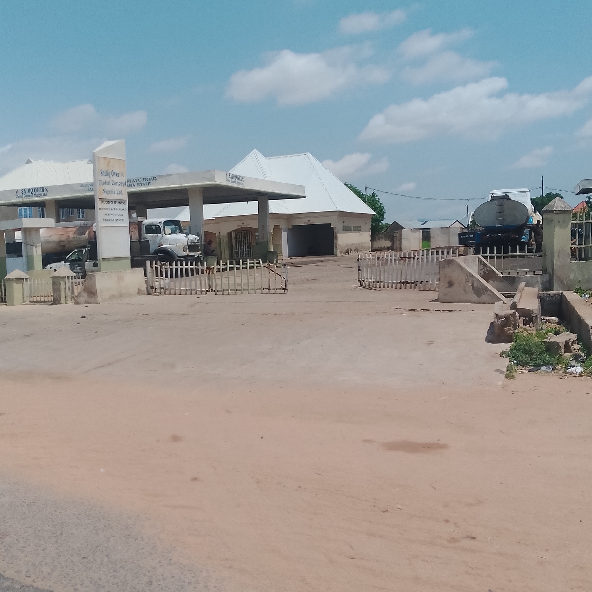 The image depicts an outdoor scene at a fuel station in Jalingo, Nigeria, under a bright, cloudy sky. In the foreground, a light brown, dusty surface dominates, showing some tire tracks and scattered debris. A white, slatted fence runs across the mid-ground.
To the left, a white building with a sloped roof is visible, with a sign that reads "SADIO OVER Global Concept Nigeria Ltd." Adjacent to this, a white truck with a fuel tanker is parked under a white canopy. The truck is white and appears to be a tanker truck.
To the right of the truck and canopy, a larger white building with a pitched roof and several windows is situated. Further to the right, another white slatted fence is present, leading towards a concrete structure with a staircase.
In the background, several other buildings with white roofs are visible, along with trees and utility poles. A man in a dark shirt is standing near the tanker truck. Another person is partially visible behind the fence on the far right. The overall environment suggests a commercial area in Jalingo, likely a fuel station serving the community.