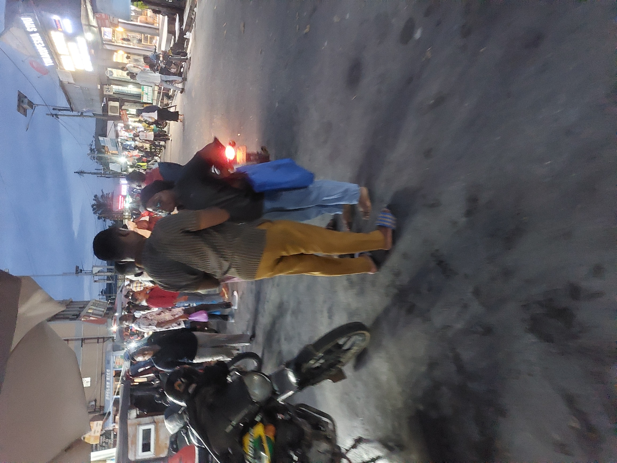 The image depicts a street scene at dusk in Dar es-Salaam, Tanzania. Two individuals stand near a motorcycle parked on the street. One person is wearing a striped shirt and mustard-colored pants, while the other is wearing a dark shirt and light-colored pants. The motorcycle is black with a round headlight. A crowd of people and shops line the street, illuminated by artificial lights. The sky is a gradient of blues and grays, indicating twilight.