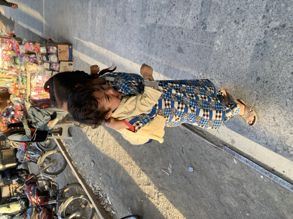 The image depicts an outdoor street scene in Quetta City Tehsil, Pakistan, under natural daylight with prominent shadows. In the foreground, two children are the primary subjects. An older child, seen from the back, wears a blue and white checkered/patchwork shalwar kameez and brown sandals. This child is carrying a younger child on their back. The younger child wears a light beige long-sleeved shirt, has dark curly hair, and is observed crying with eyes squeezed shut and mouth open. The younger child's arms are around the older child's neck/shoulders.
In the mid-ground, to the left of the children, multiple motorcycles are parked on a rough, unpaved dirt and gravel shoulder containing scattered debris. Further into the mid-ground, an adult male with a beard is seated on a light-colored stool, wearing a dark vest over a light shirt and traditional loose trousers. He is looking down at his hands. Behind him and extending to the right is a street vendor stall displaying a variety of colorful items, including toys and packaged goods, arranged on shelves and within boxes.
The ground surface on the left is unpaved and irregular, while the area occupied by the children and extending to the right is a smooth, grey paved surface with a faint white line visible. A concrete curb separates the unpaved and paved sections. Additional partial figures are visible in the far background.