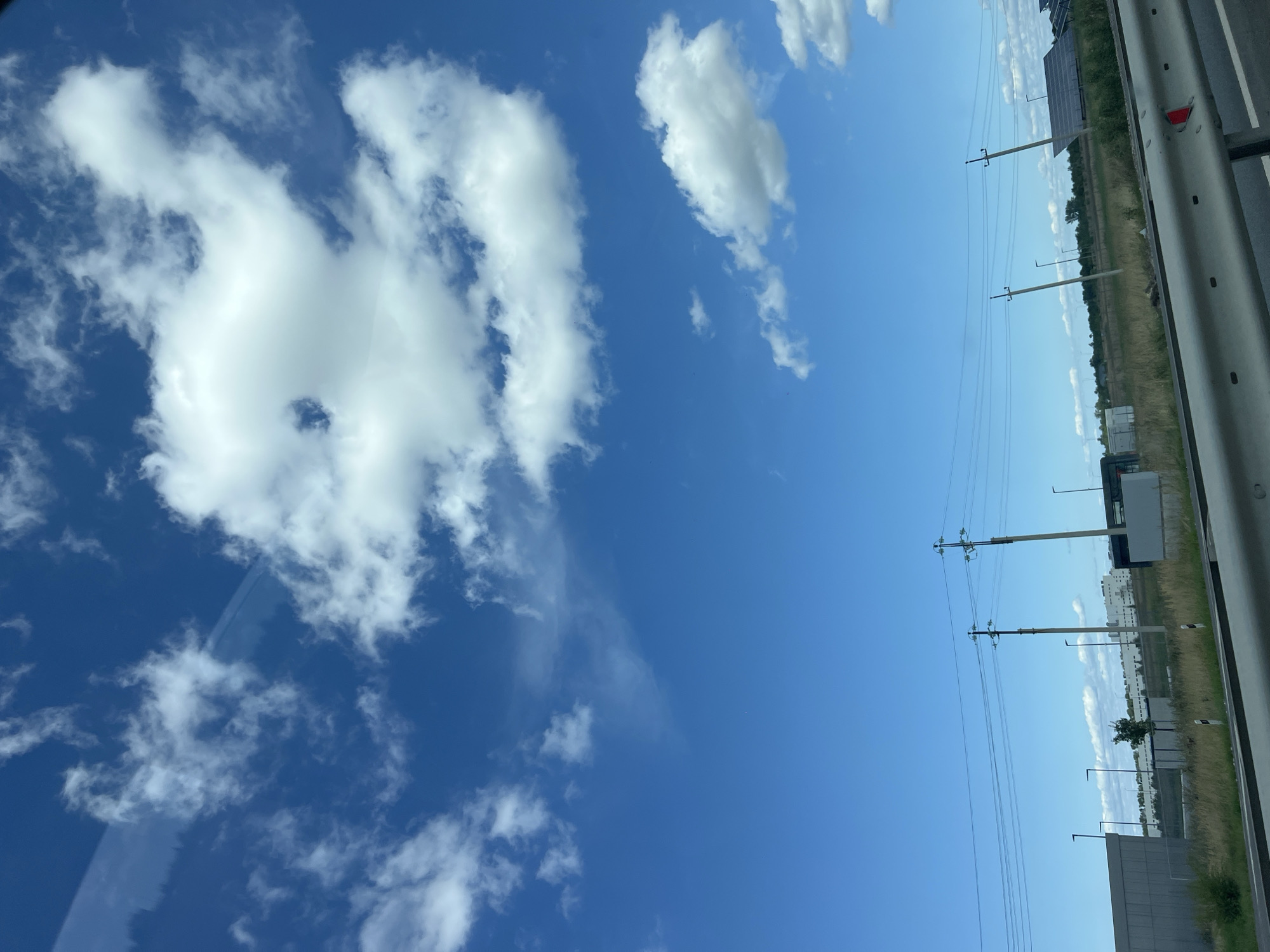 The image depicts a bright, clear blue sky with scattered white clouds. In the foreground, a metal guardrail is visible, suggesting a view from a vehicle or overpass. Beyond the guardrail, a grassy area transitions into a landscape with several power poles and overhead wires. In the background, there are some industrial-looking buildings and solar panels. The overall scene is typical of an outdoor environment, possibly along a highway or a suburban area. There are no people visible in the image. The location is not identifiable as Ulyanovsk, Russia.