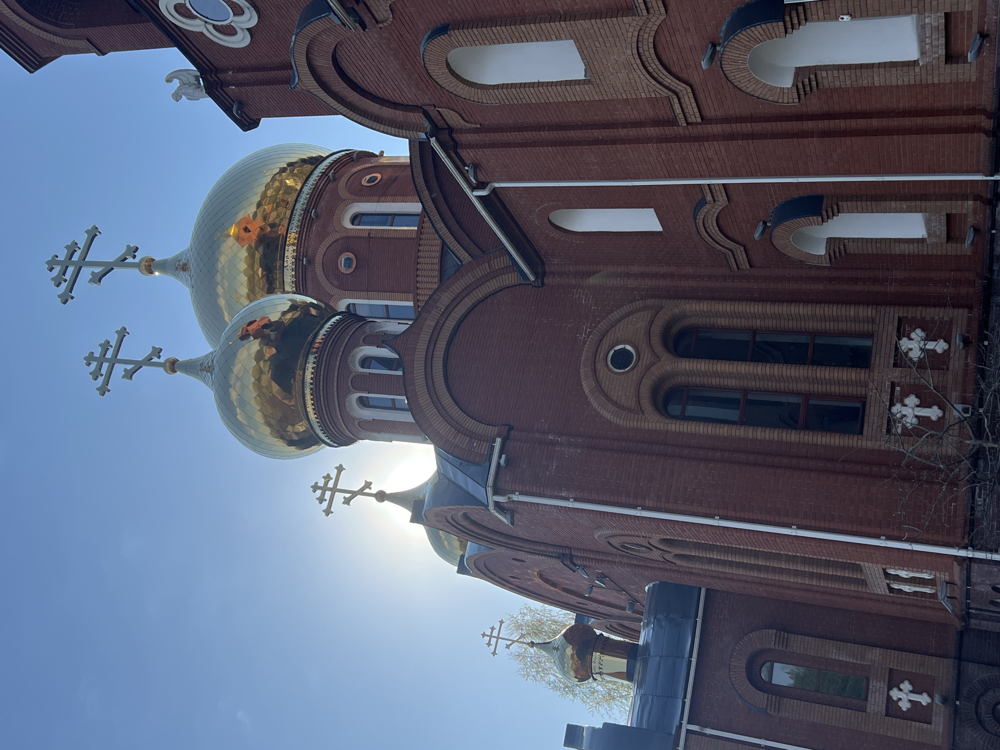 The image depicts the exterior of a brick Orthodox Church in Chaikovsky, Russia, under a bright blue sky. The most prominent features are the onion domes, covered in golden-colored tiles, with Orthodox crosses atop each dome. The sun is shining brightly, causing some glare. The church is made of red brick. The image includes architectural details such as arched windows and decorative brickwork. There is a tree branch visible on the right side of the frame.