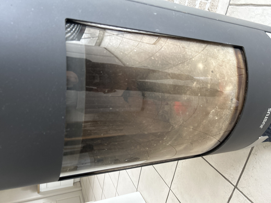 An interior scene captured in Veauchette, France, depicts a dark grey, modern, cylindrical "JUSTUS" brand wood-burning stove positioned on a light-colored tiled floor with grey grout lines. The stove's prominent glass viewing panel is significantly smudged and dusty. Through the soiled glass, a residual layer of ash and partially combusted material is visible within the combustion chamber, with a faint reddish glow suggesting the presence of embers or residual heat at the lower-middle portion. A distinct reflection of a person, identifiable by dark legwear and a lighter torso, is superimposed on the stove's glass. The reflection also shows more of the tiled floor extending away from the stove and a white cabinet or piece of furniture in the background to the left.
