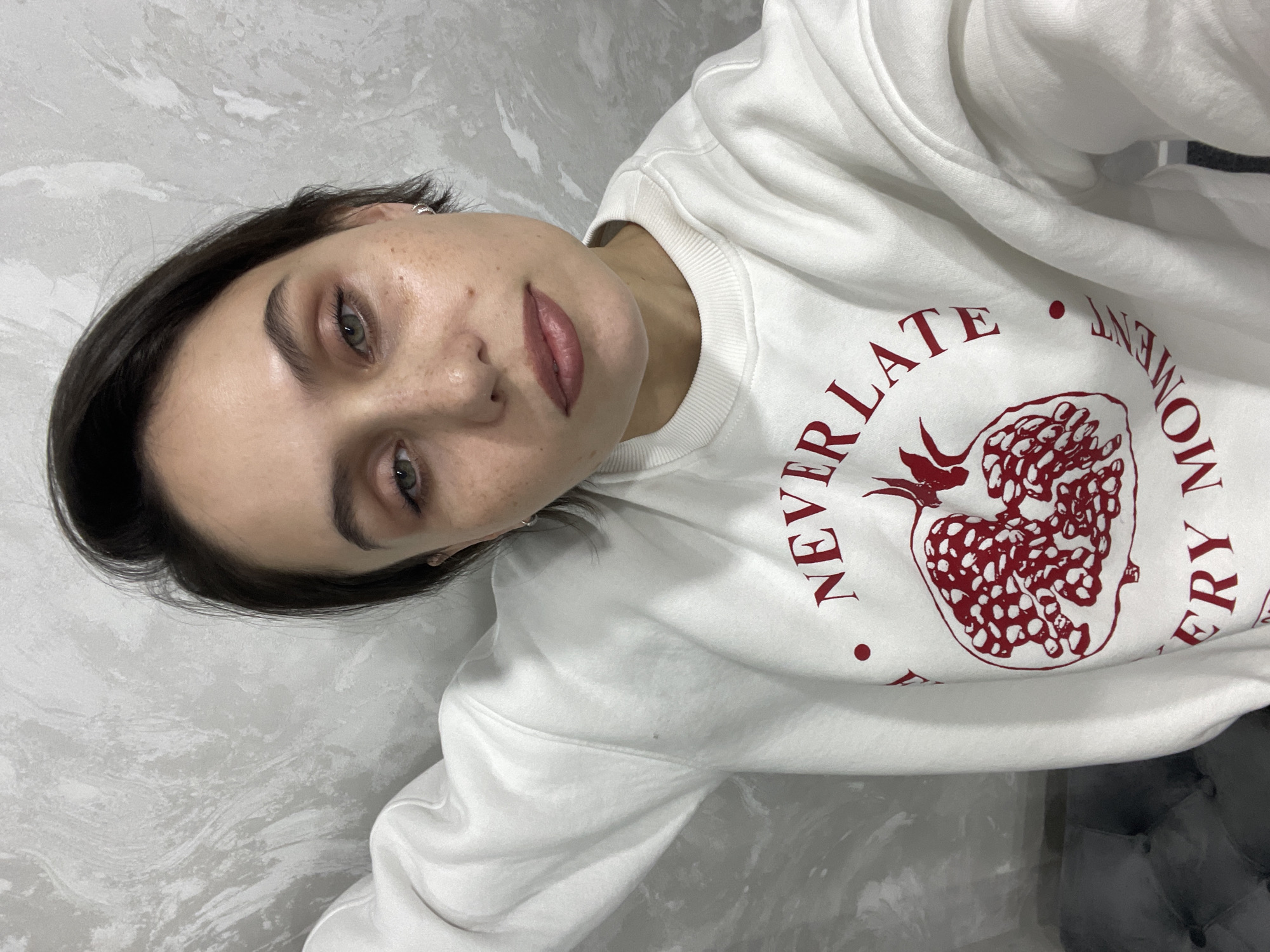 A person is wearing a white sweatshirt with a red graphic design and text reading "NEVER LATE EVERY MOMENT". The person has short, dark hair and light skin with freckles. The background is a gray textured wall. There is no evidence to suggest the location is Izhevsk, Russia.