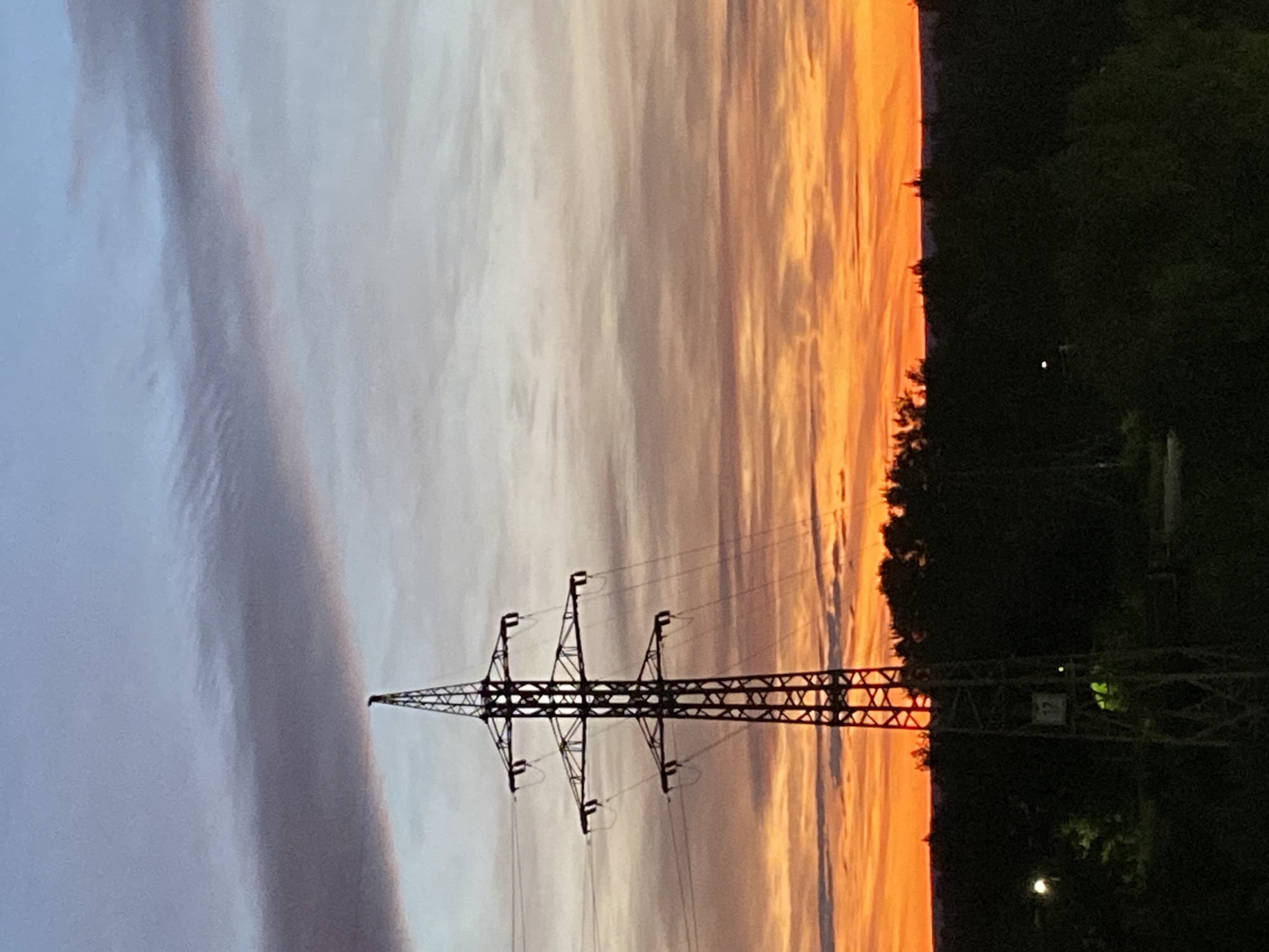 This is a vertical, medium-shot photograph of a power line pylon silhouetted against a dramatic sunset sky. The sky transitions from soft grays and whites on the left to vibrant oranges and yellows on the right, with wispy clouds streaking across. The pylon, made of dark metal lattice, dominates the center of the frame, with its long arms extending outwards to hold the power lines. The power lines themselves are thin, dark lines stretching across the sky.
To the right of the pylon, a dense line of dark green trees forms the horizon. Beyond the trees, there are a few faint lights visible, suggesting some distant human habitation. The overall scene is one of industrial infrastructure set against a natural, ephemeral event. The time of day is clearly sunset, and the dramatic colors of the sky evoke a sense of awe or perhaps the closing of a day. There is no discernible text in the image.