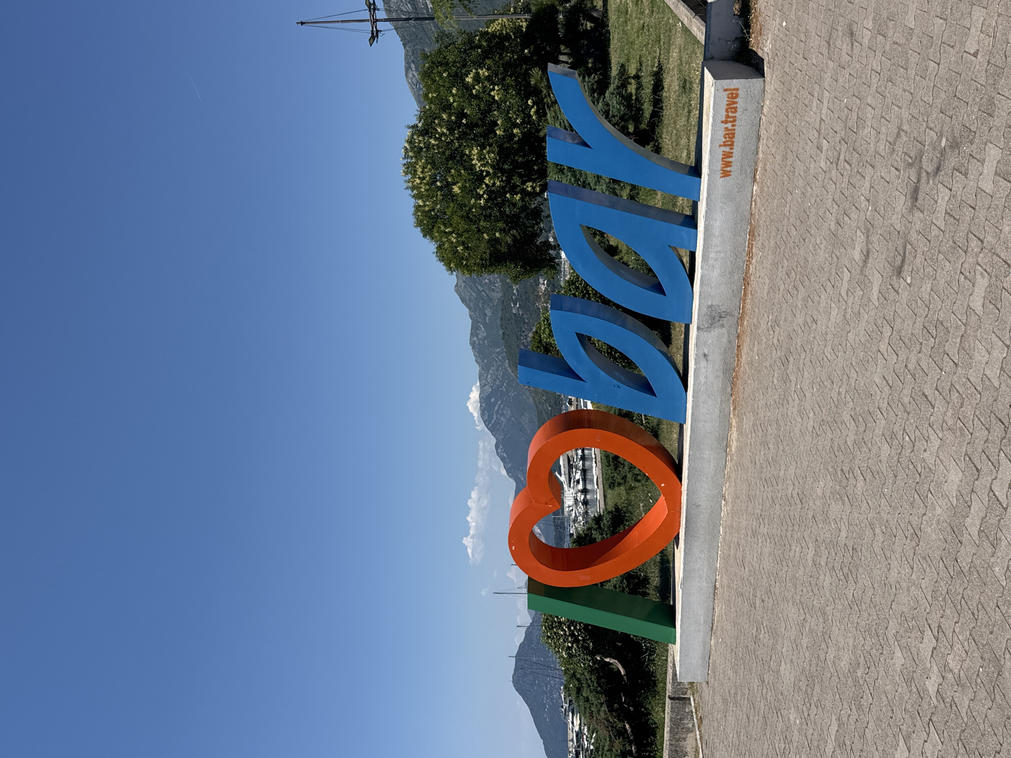 A large, colorful, 3D sign spelling out "I LOVE BAR" is displayed outdoors. The "I" is represented by a green rectangle. The "LOVE" is depicted by a large, orange heart. The letters "BAR" are rendered in blue with a distinctive, stylized font. The sign is situated on a gray concrete base, with the website "www.bar.travel" printed in orange on the right side of the base. The background reveals a scenic view of a bay dotted with sailboats and yachts, lush green trees, and rolling mountains under a clear blue sky. The foreground is paved with gray interlocking bricks.